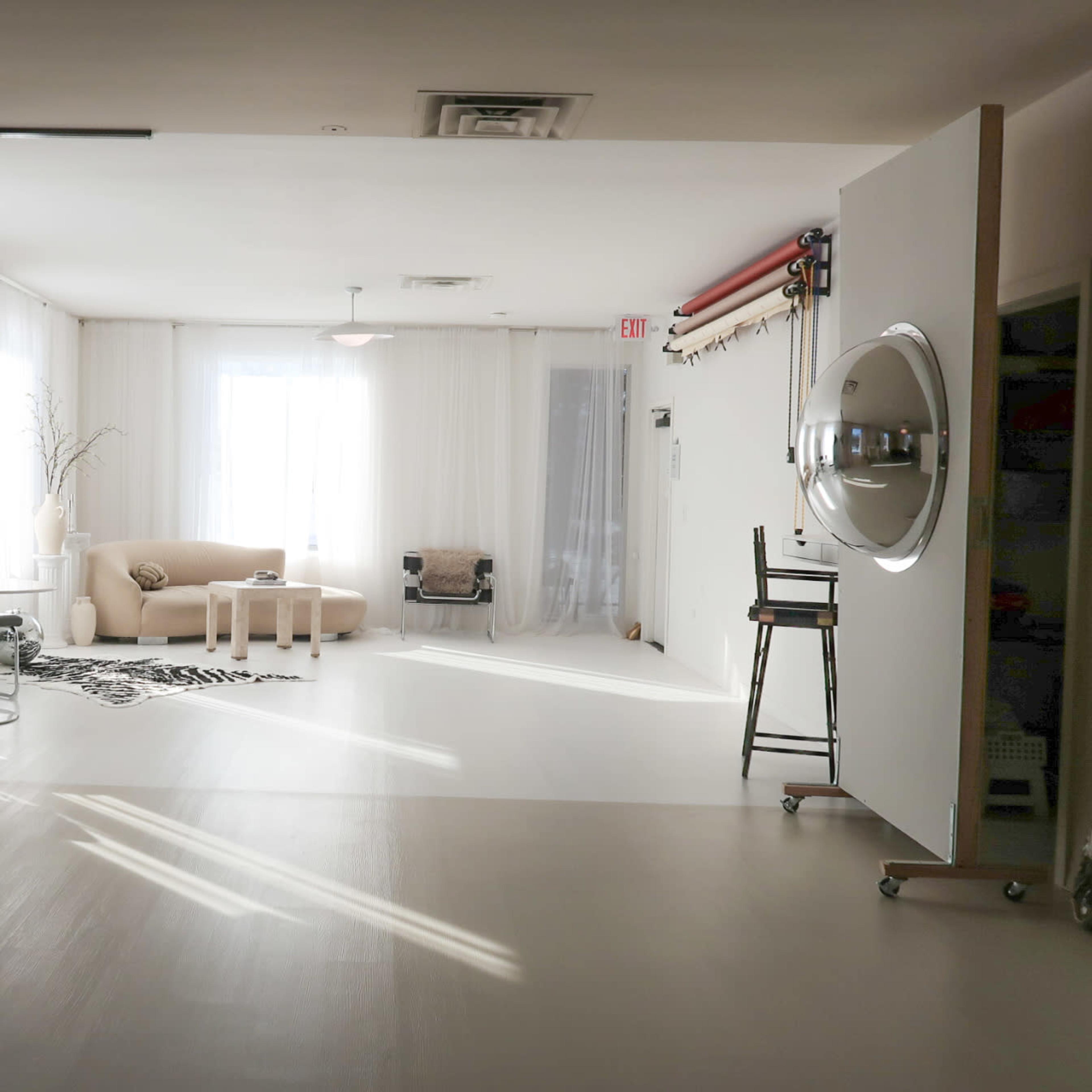 A bright studio space features large windows, a couch, a small table, and photography equipment, with white walls and a light-colored floor.