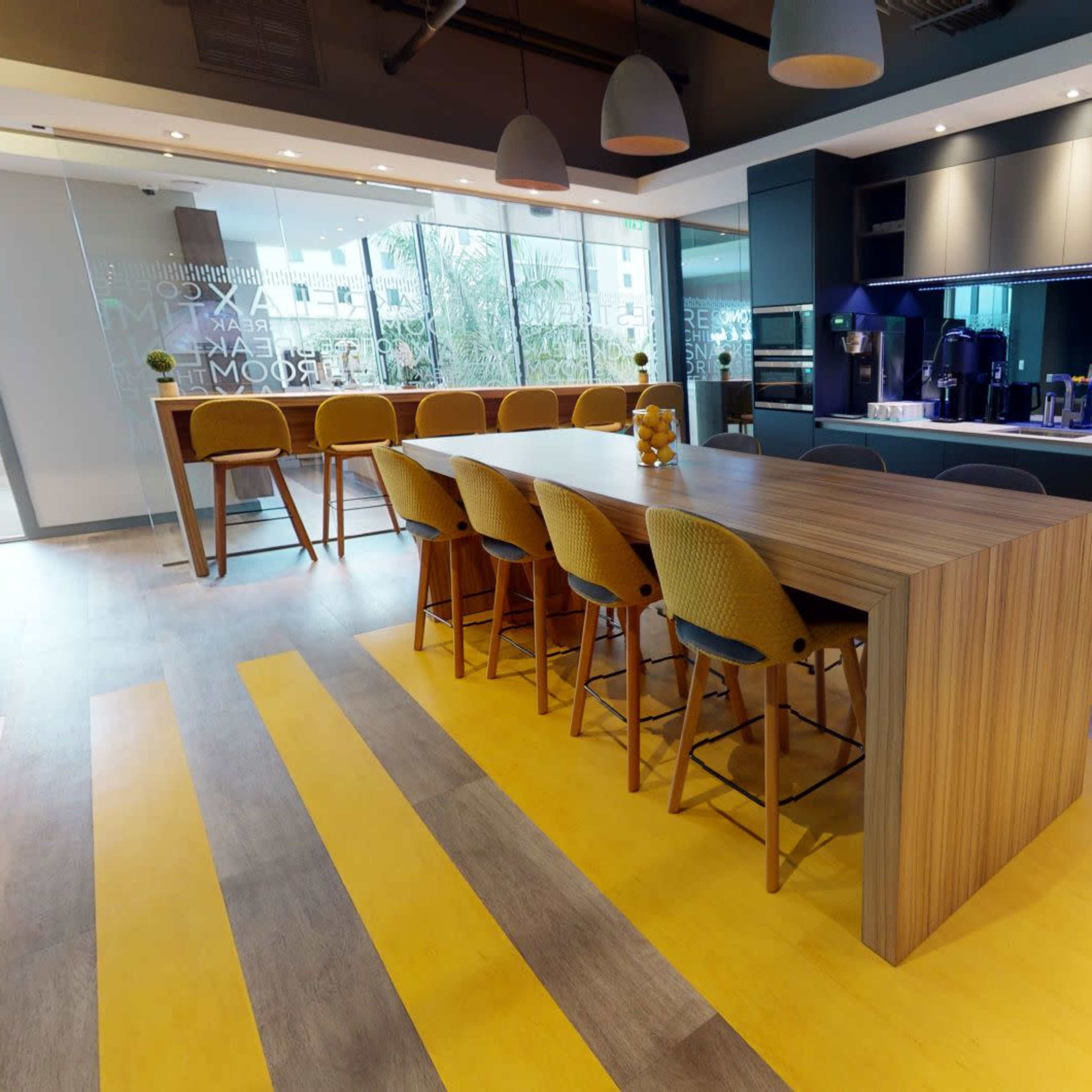 A modern kitchen area with a long wooden bar, high stools, various appliances, and a bright yellow and gray floor design.