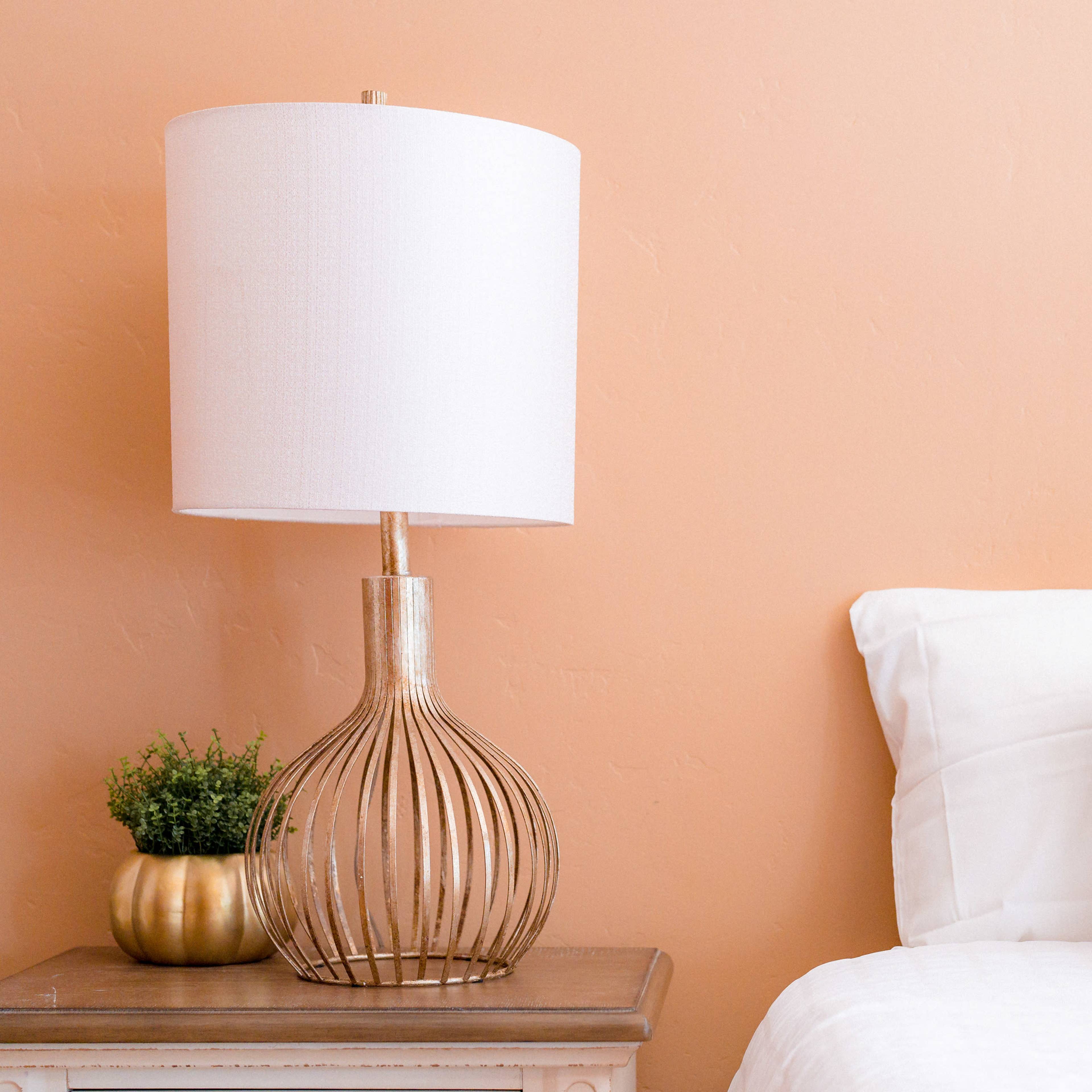 A lamp with a white shade is placed on a wooden side table next to a neatly made bed against a peach-colored wall.