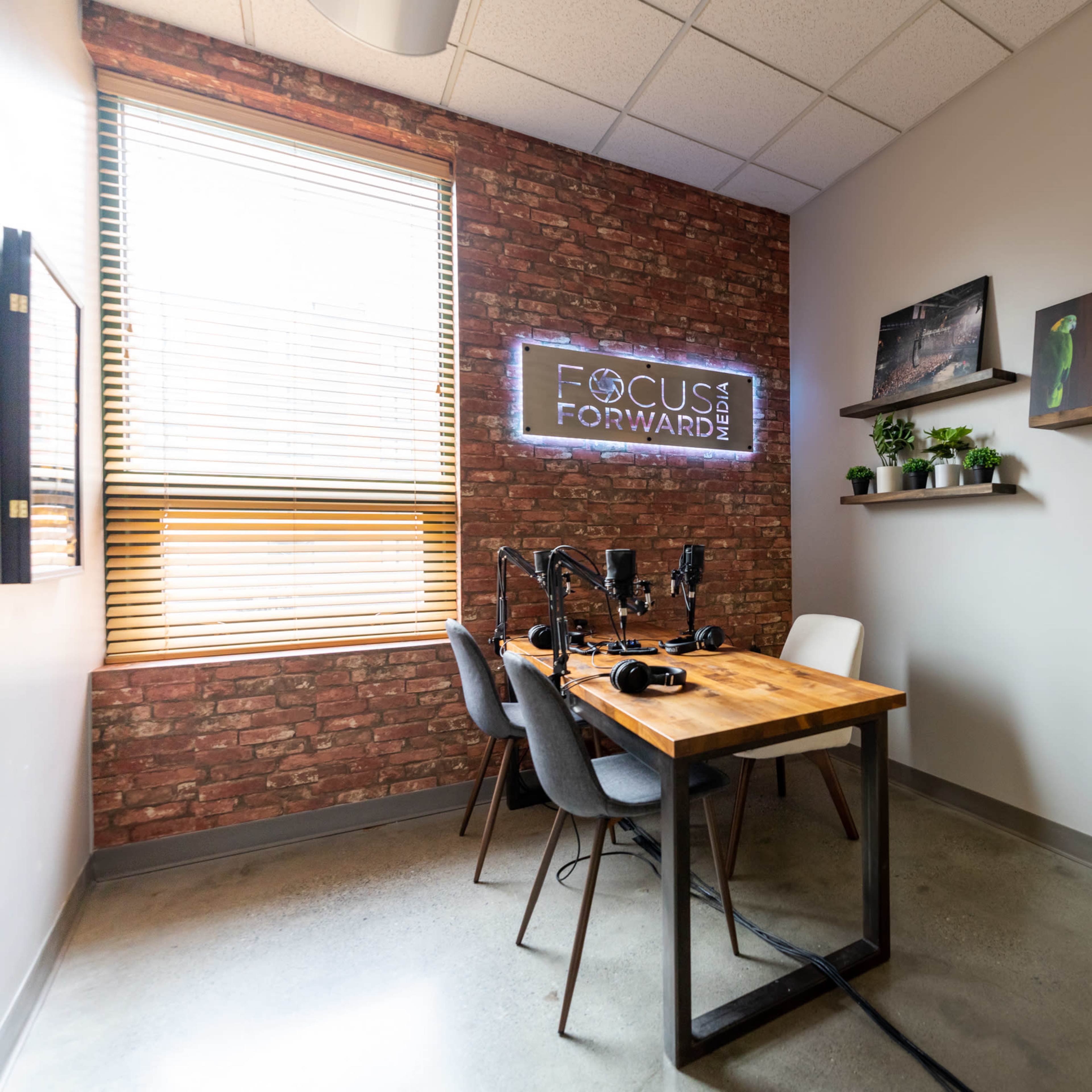 A small recording studio featuring a wooden table with microphones, chairs, a brick wall, and a door labeled "STUDIO."