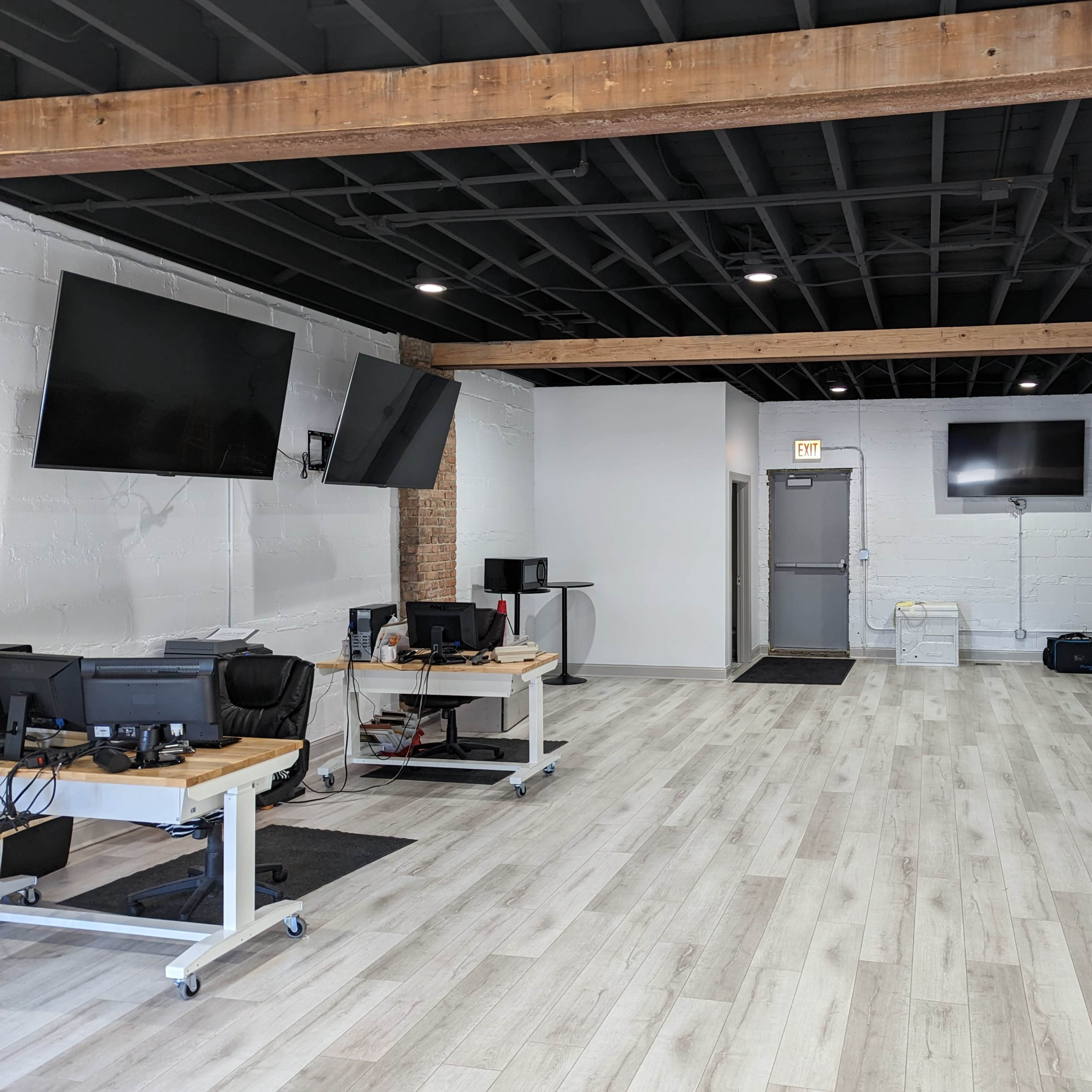 An open office space with multiple workstations, two large wall-mounted screens, and a door leading to another area.