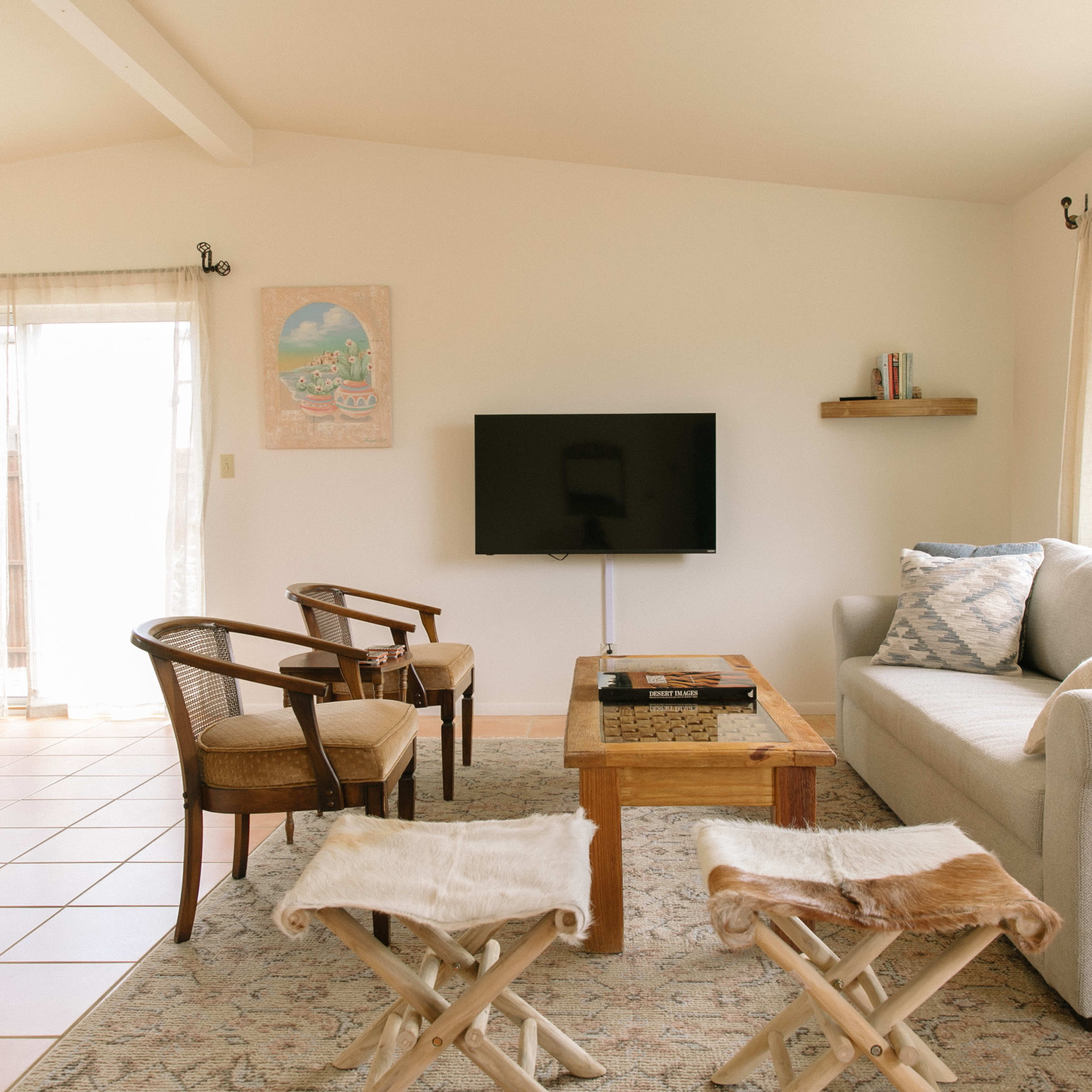 A bright living room featuring a gray sofa, a wooden coffee table, two chairs, and a television mounted on the wall, with a view of a dining area and large windows.