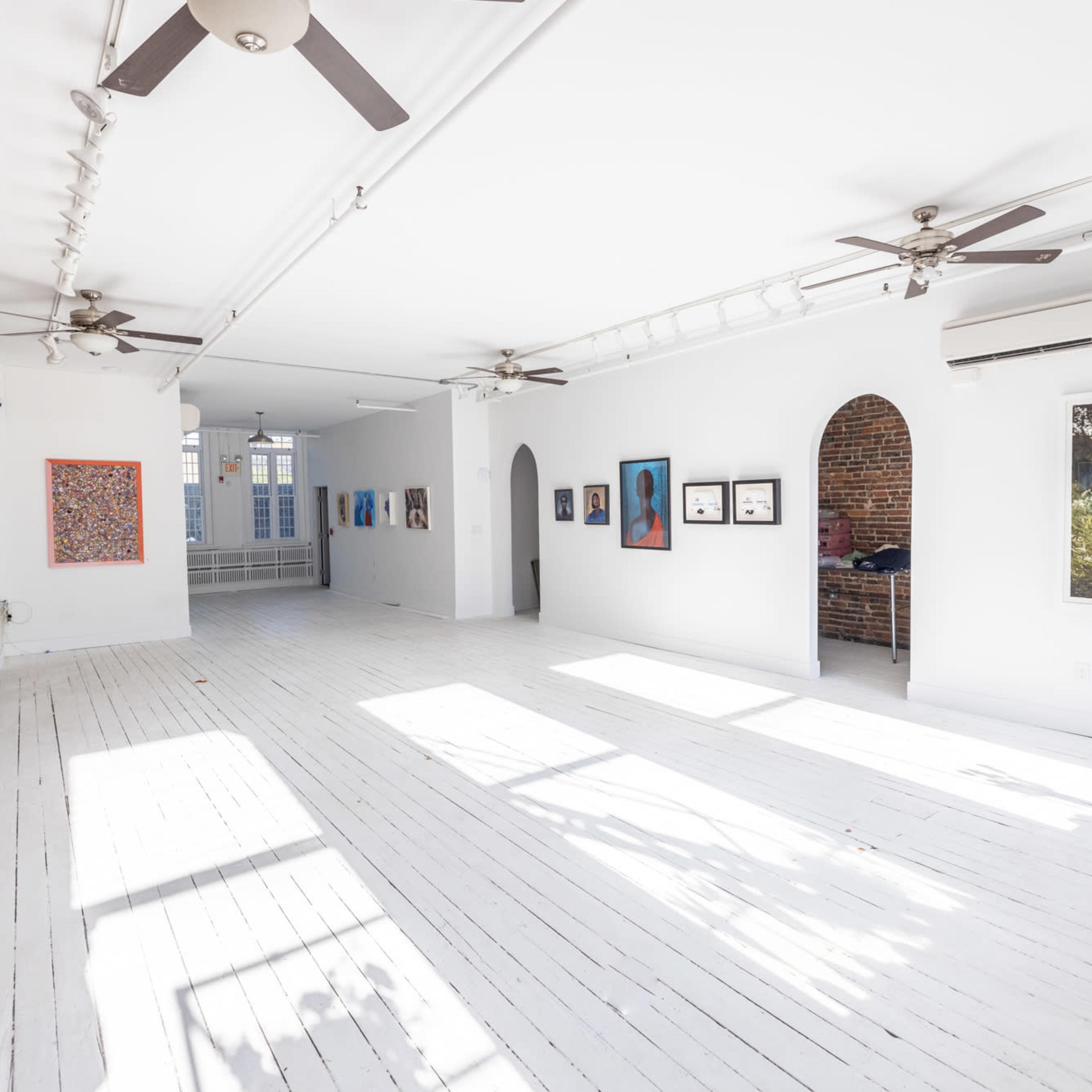 A spacious art gallery with white wooden floors, exposed brick walls, and various framed artworks hung on the walls.