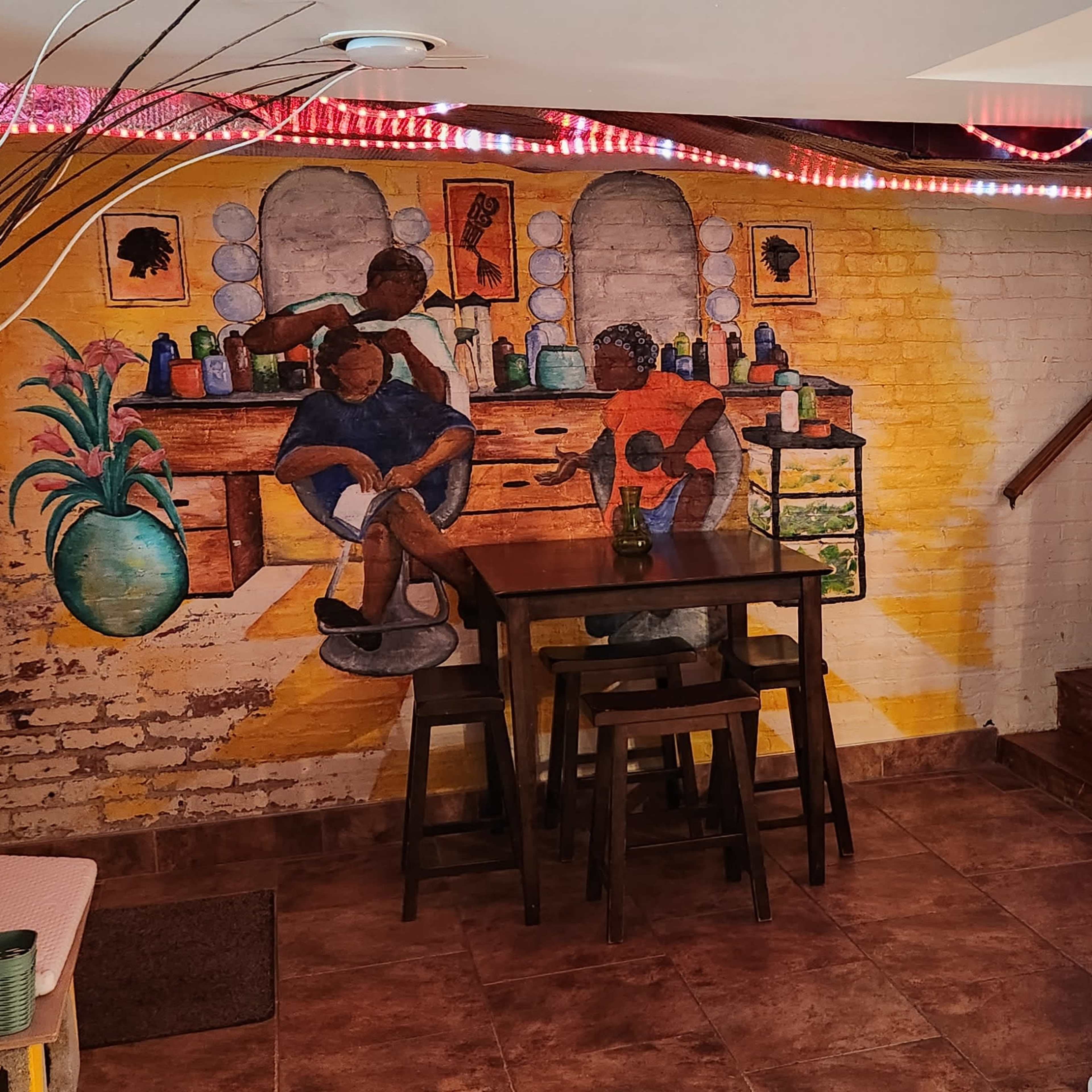 A basement interior featuring a mural of two people at a countertop, a wooden table with chairs, and decorative lighting along the ceiling.