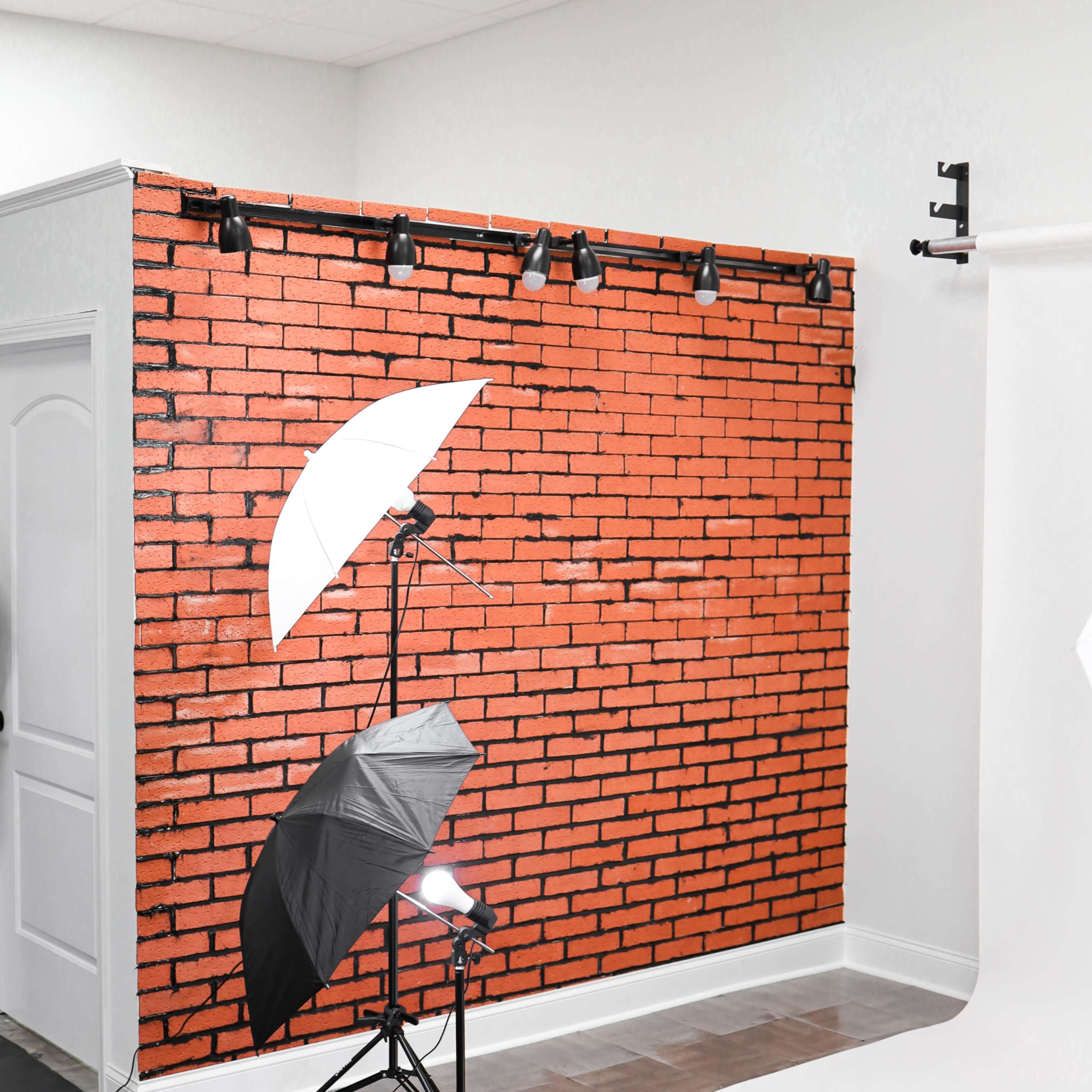 A photography studio with a red brick backdrop, lighting equipment, and a white seamless backdrop.