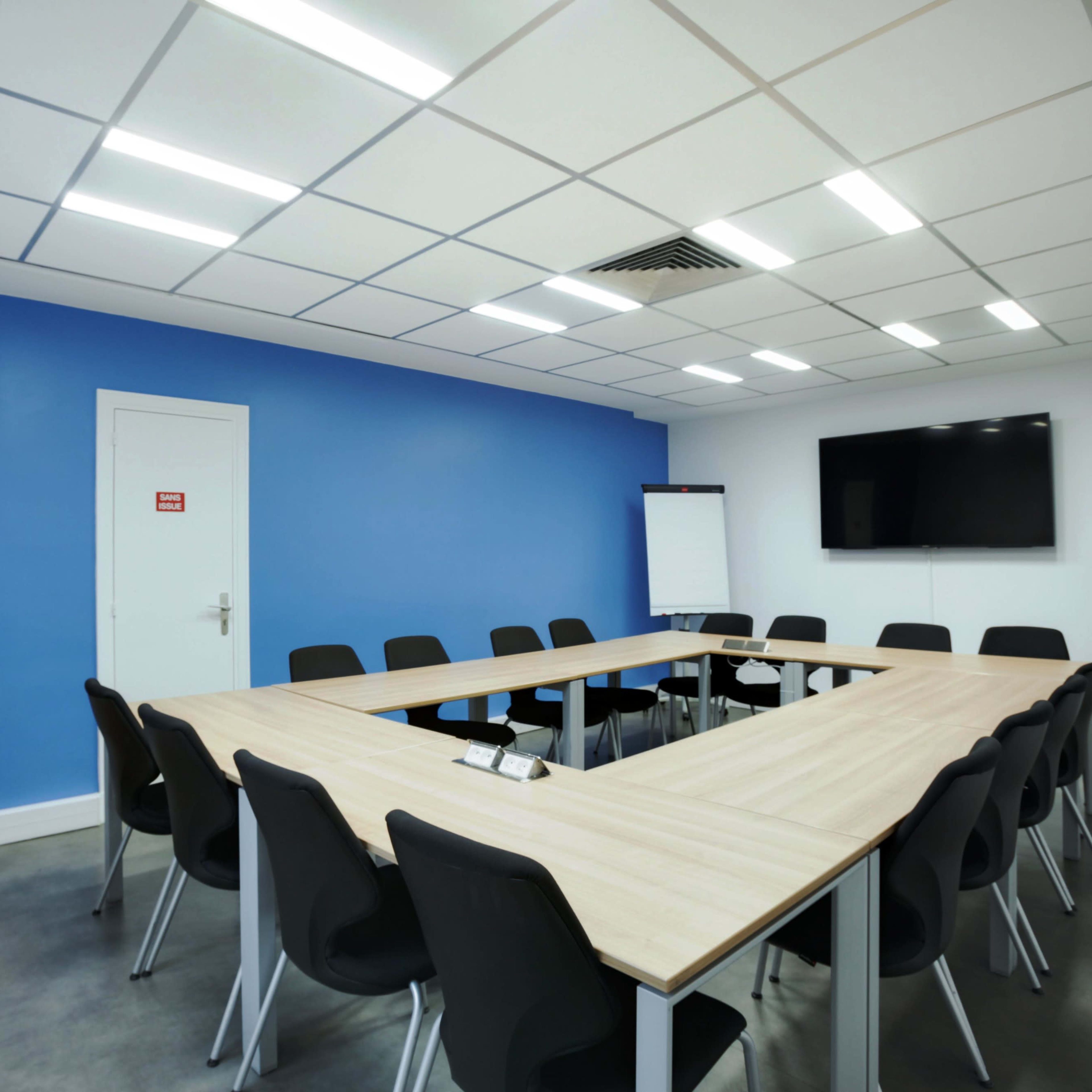 A modern conference room featuring a large wooden table arranged in a U-shape, surrounded by black chairs, with a blue accent wall and a wall-mounted TV.