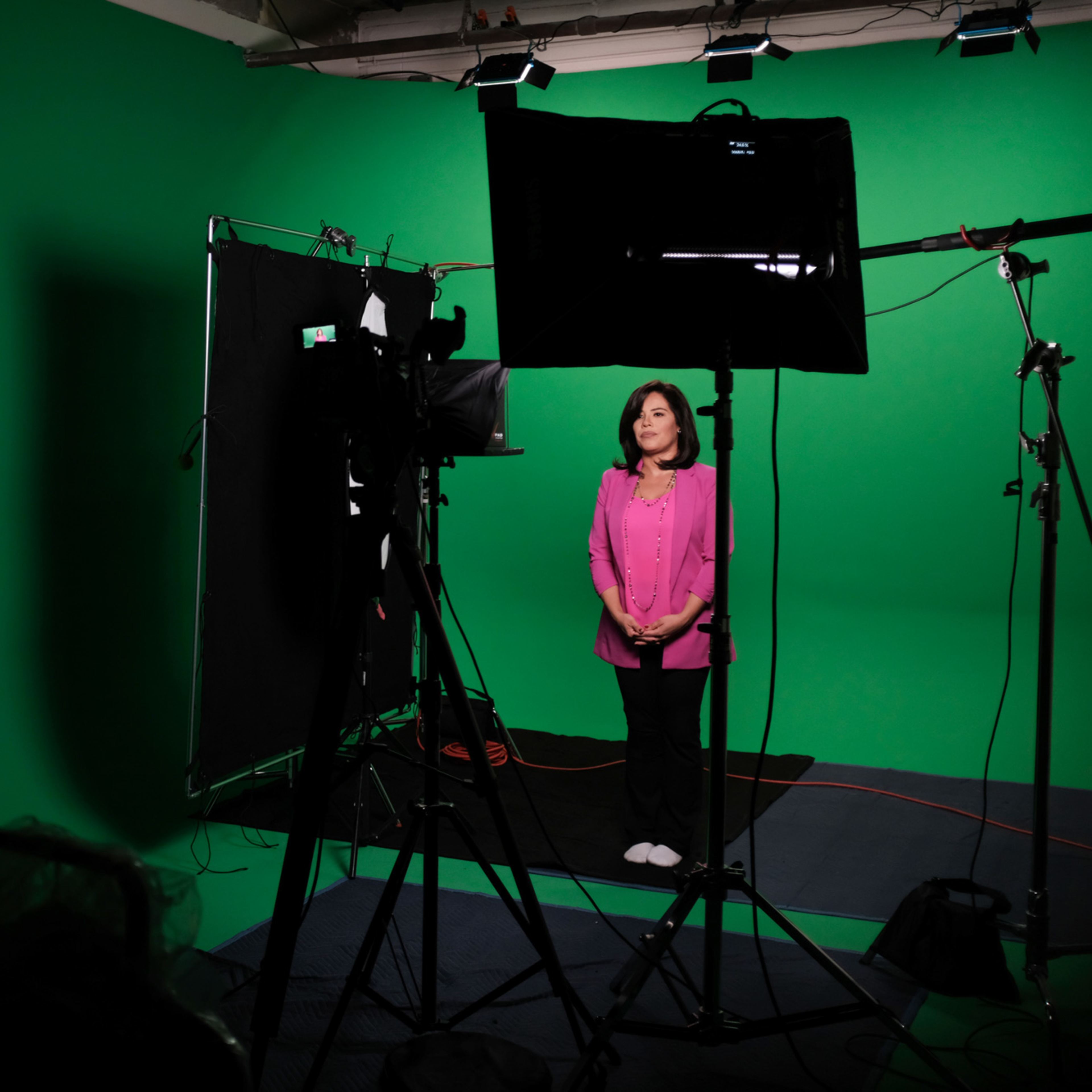 A woman in a pink top stands in front of a green screen while being filmed, with camera equipment and a monitor positioned nearby.