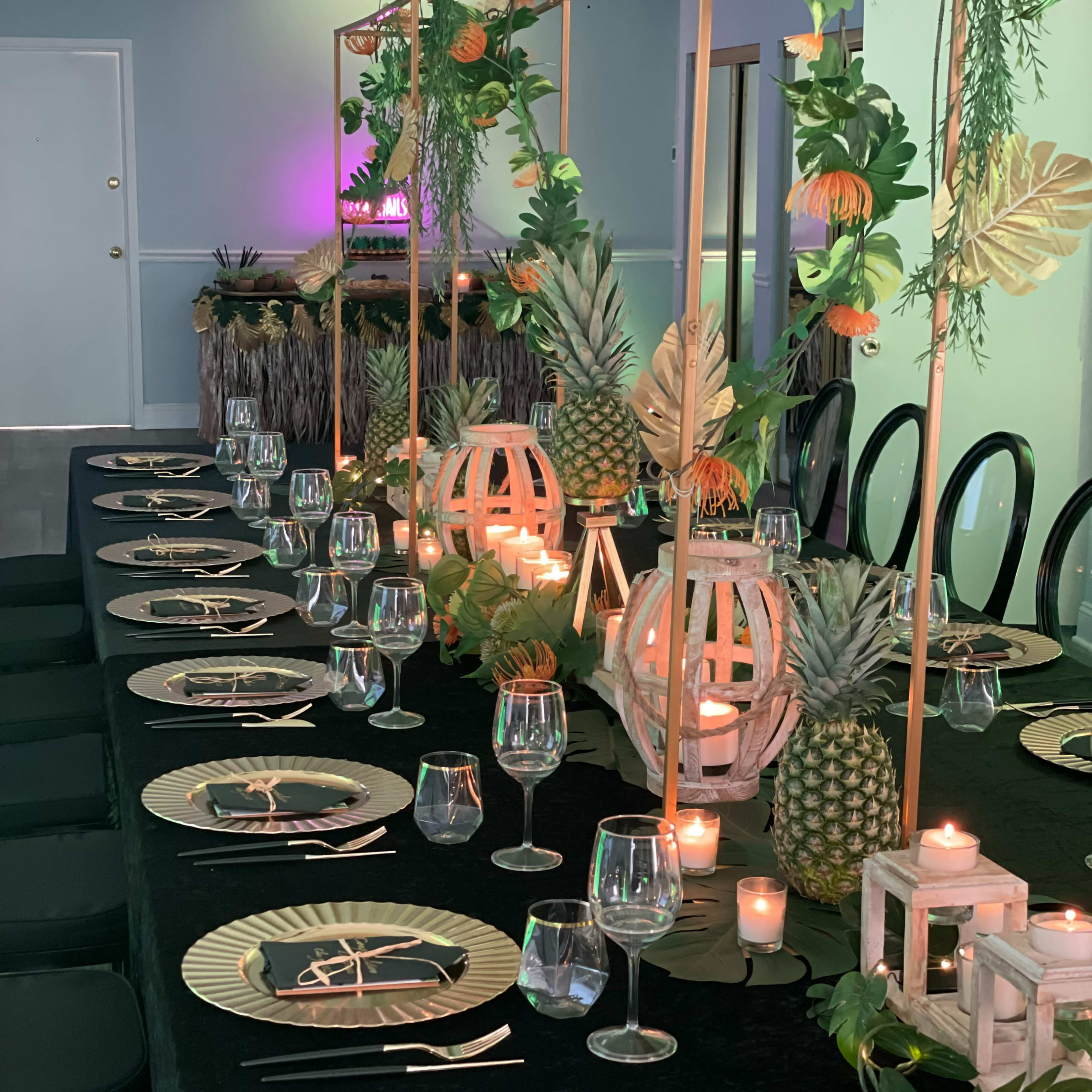 A long, elegantly set dining table is adorned with tropical decorations, including pineapples and lanterns, along with candles and glassware.