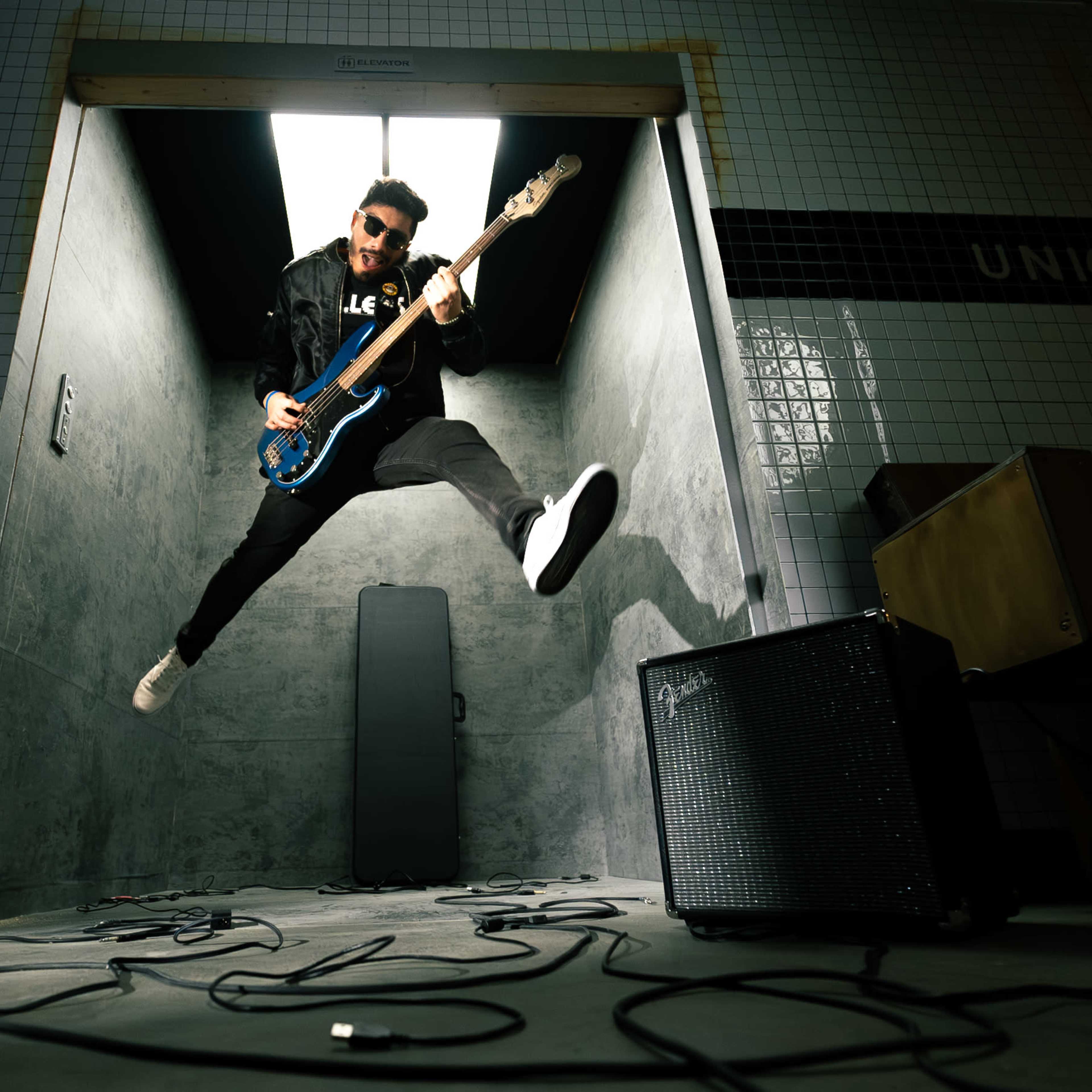 A musician jumps in mid-air while playing a blue bass guitar in a dimly lit room with tiled walls.