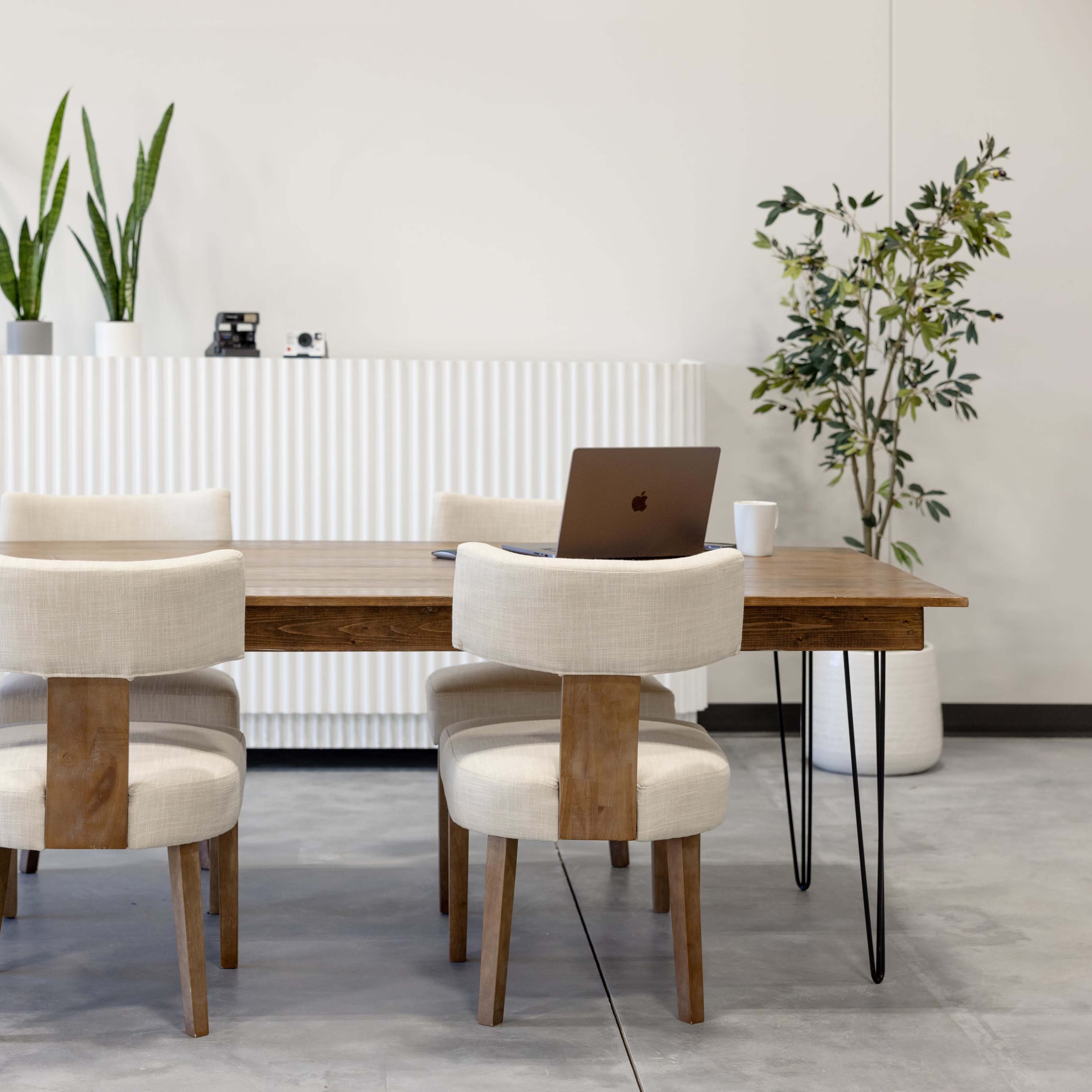 A wooden dining table with four chairs is positioned in a minimalist room, accompanied by a laptop and potted plants against a white wall.