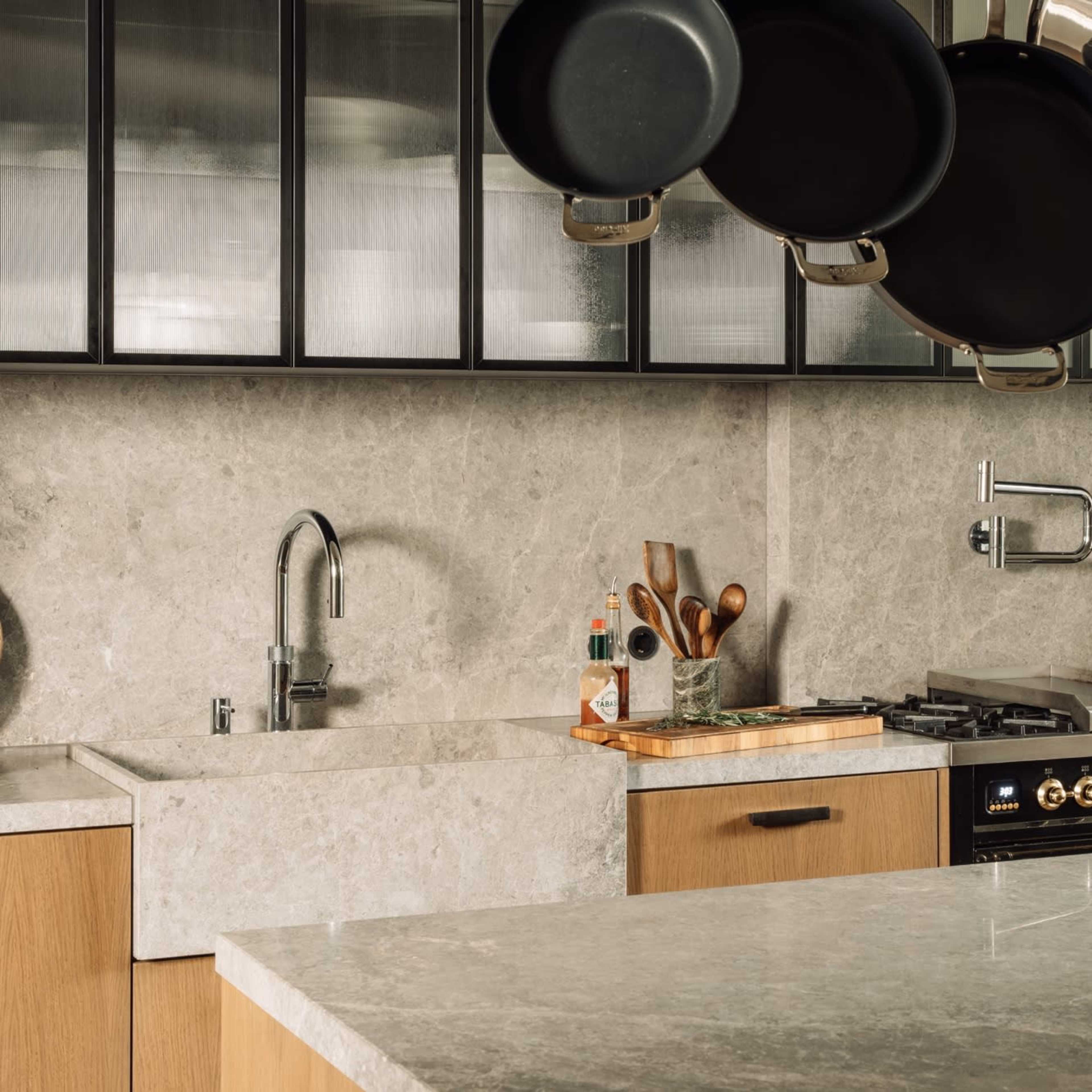 A modern kitchen features gray stone surfaces, wooden cabinetry, and hanging pots above a gas stove.