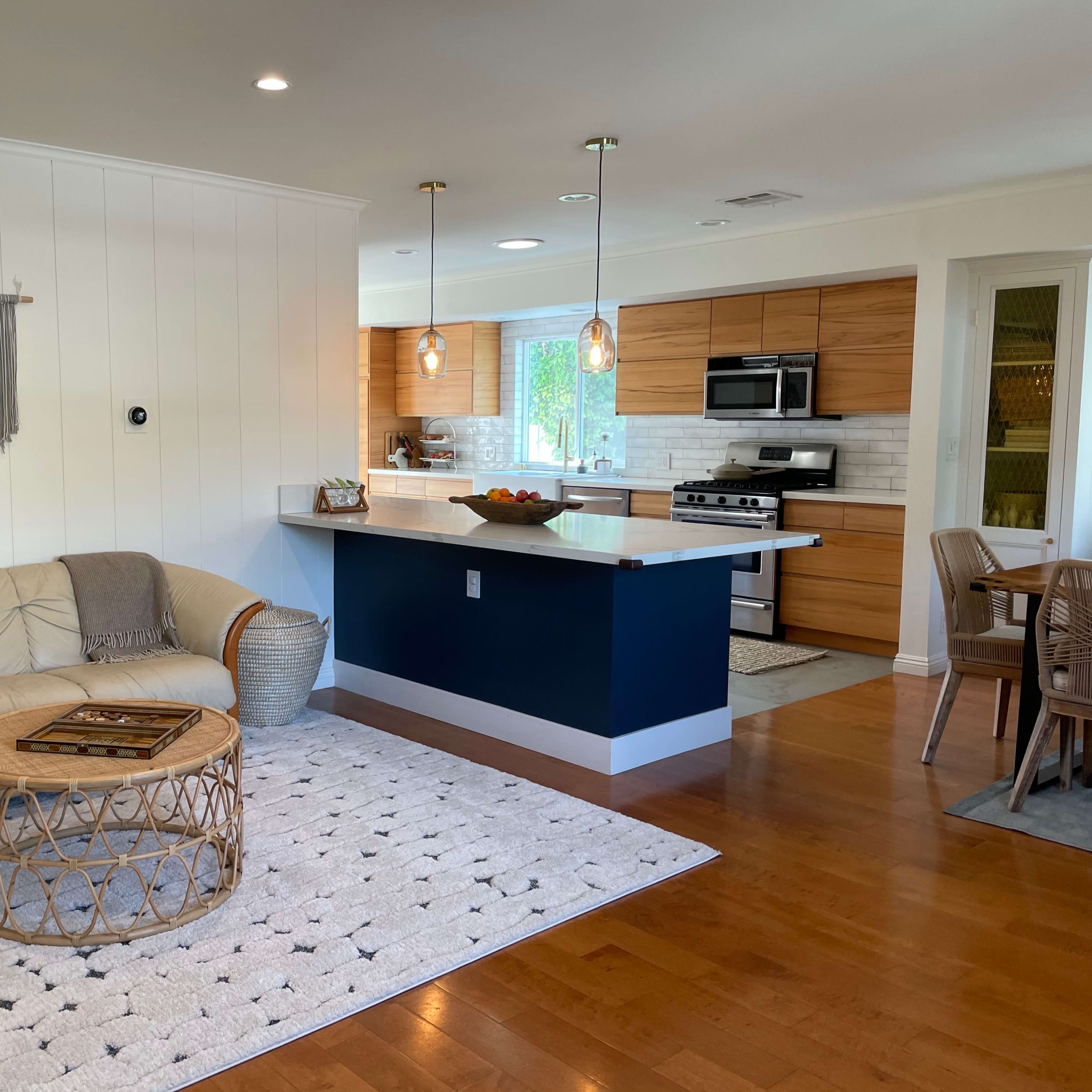 A modern living area features a light-colored sofa, a round woven coffee table, and an open kitchen with wooden cabinetry.
