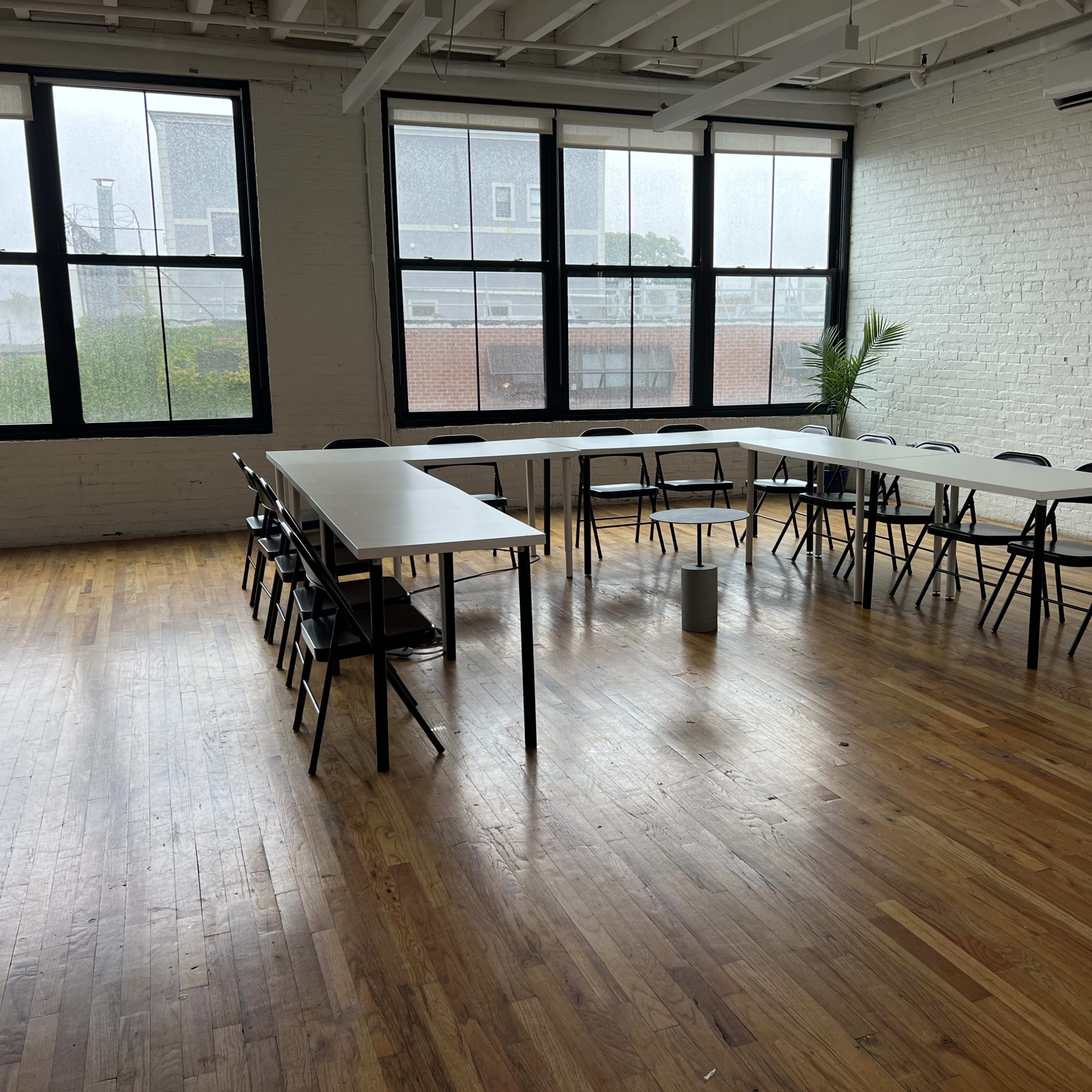 A large, rectangular table is surrounded by black folding chairs in a spacious room featuring large windows and wooden flooring.
