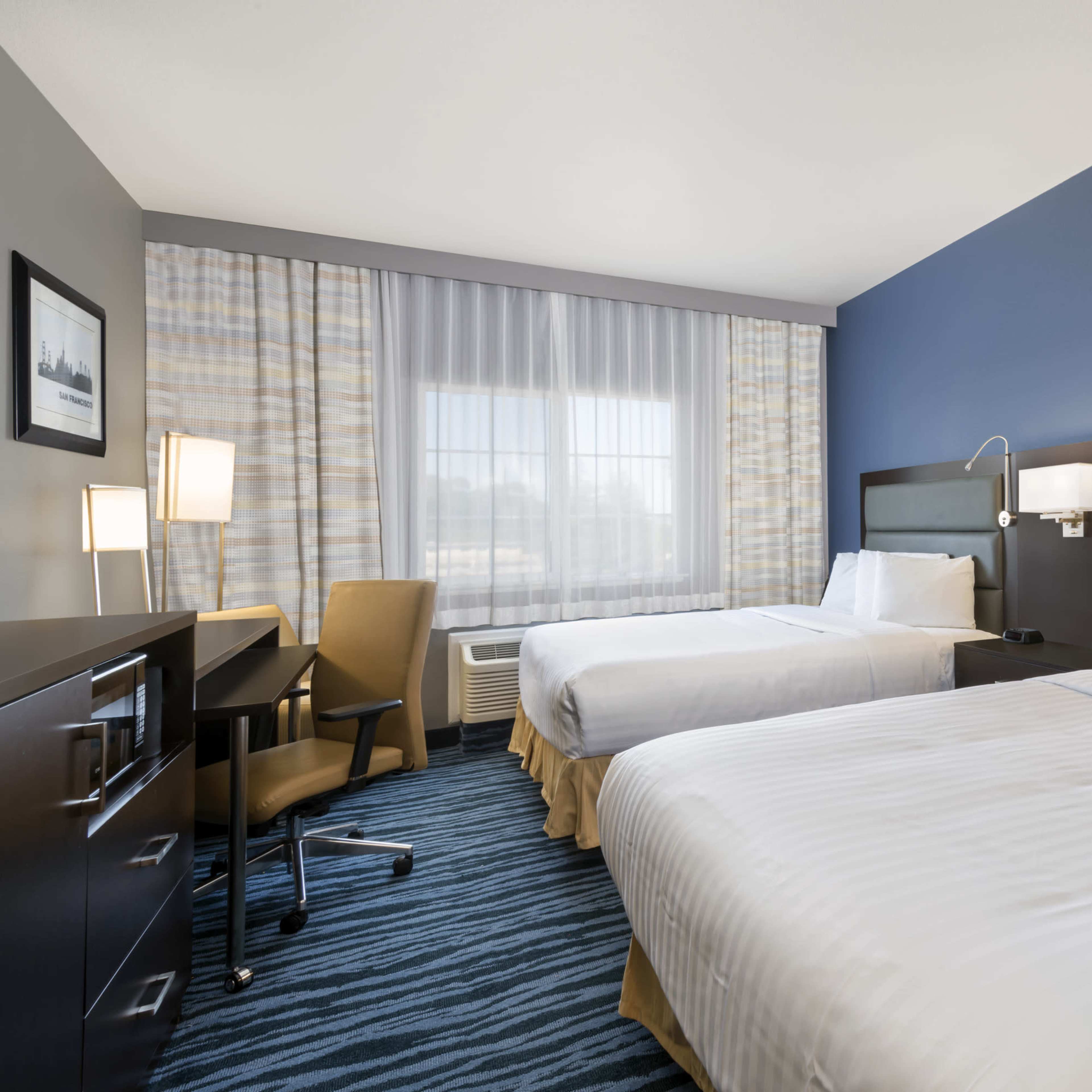 A hotel room with two beds, a desk, and a chair, featuring a large window with sheer curtains.