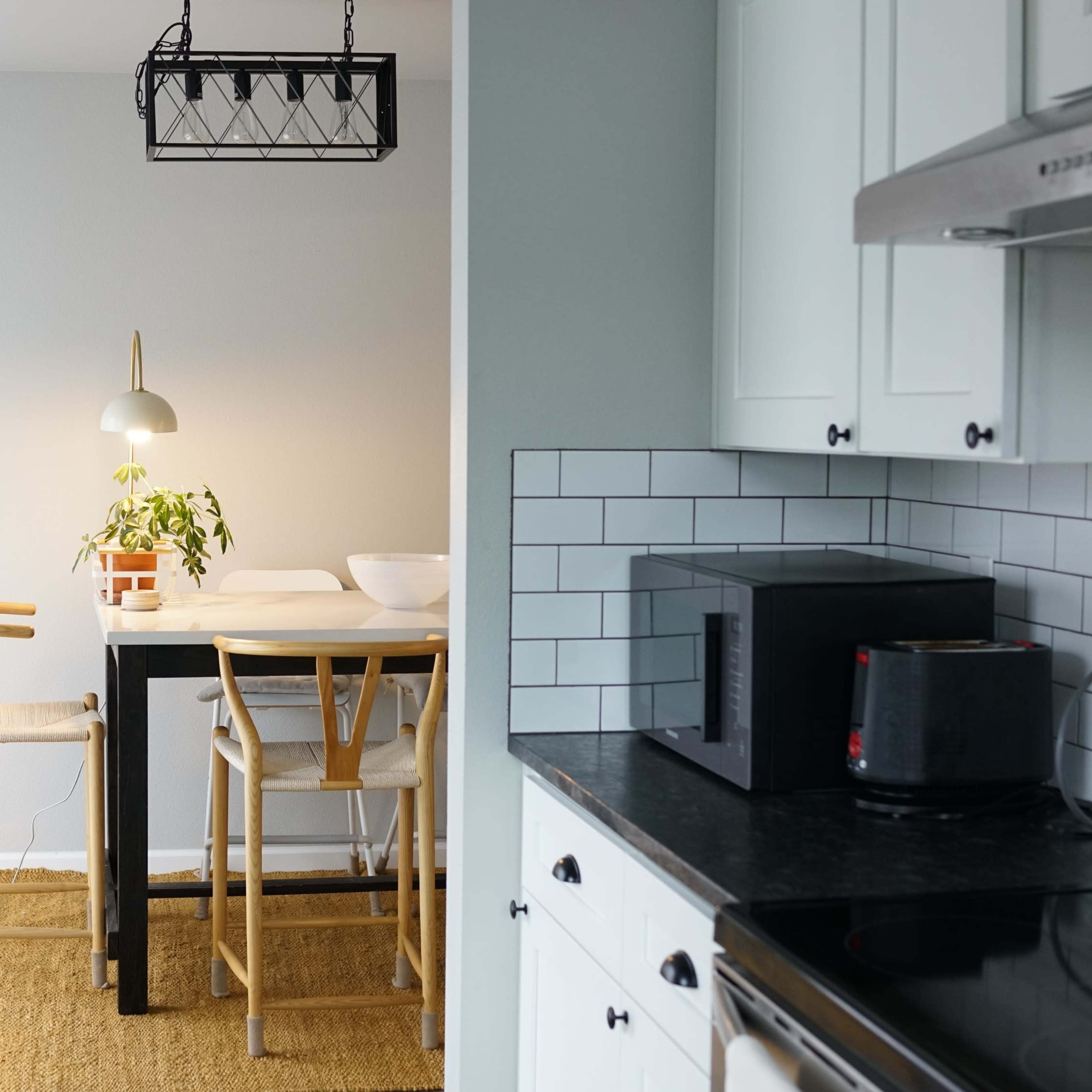 A modern kitchen opens into a dining area featuring a simple table with high stools and a potted plant.
