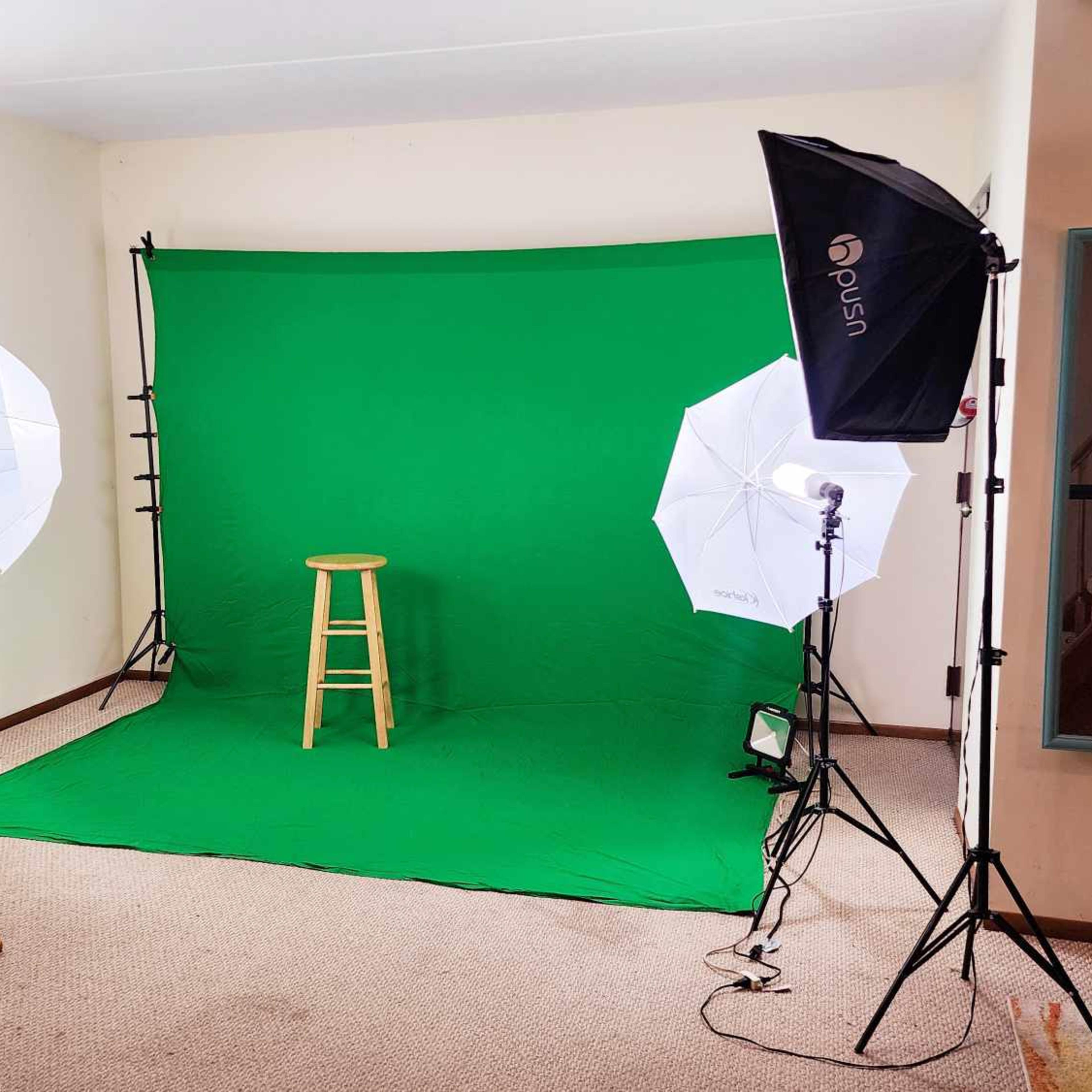 A photography studio is set up with a green backdrop, two softbox lights, and a wooden stool positioned in front.