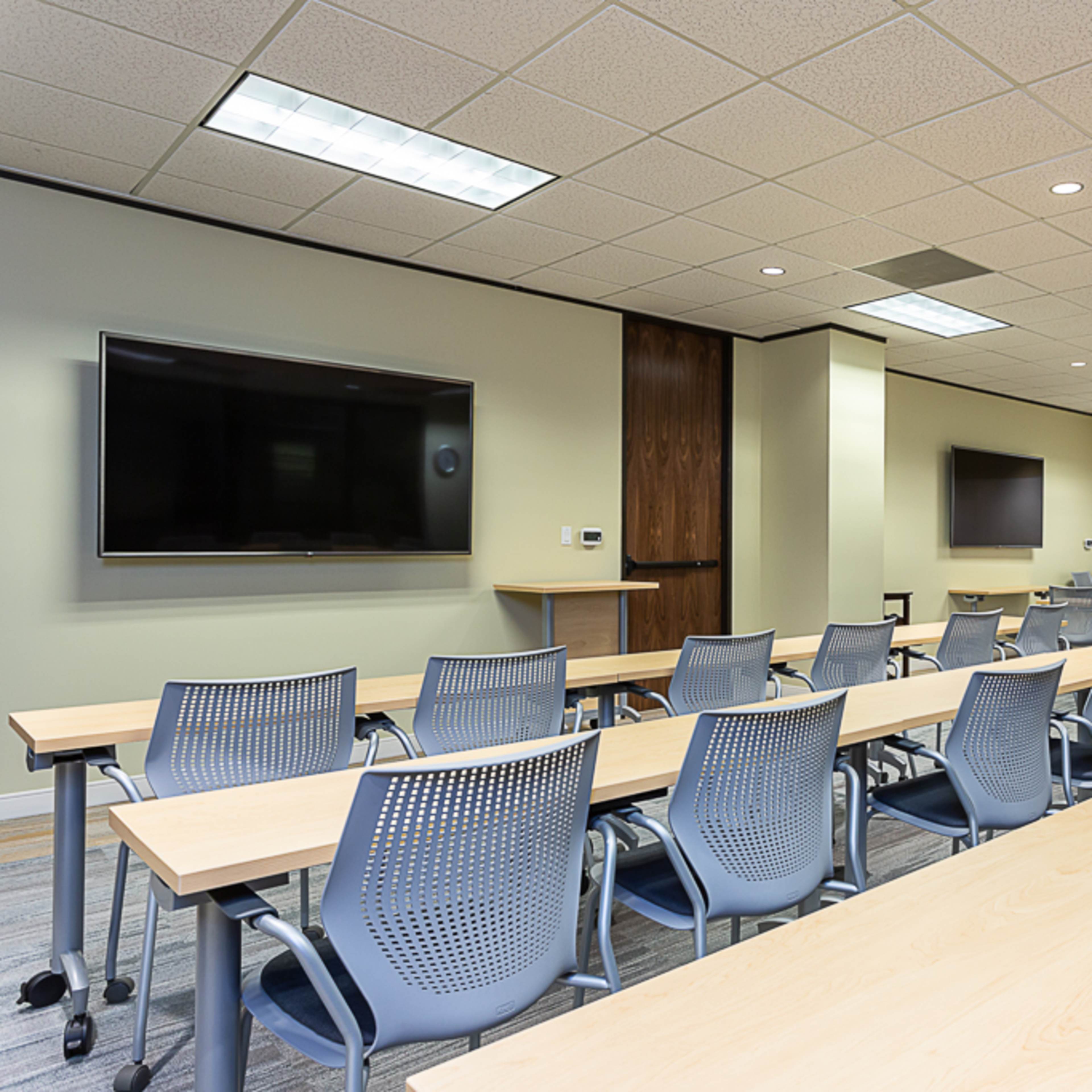 A spacious classroom setup with rows of tables and chairs, large wall-mounted screens, and a whiteboard.