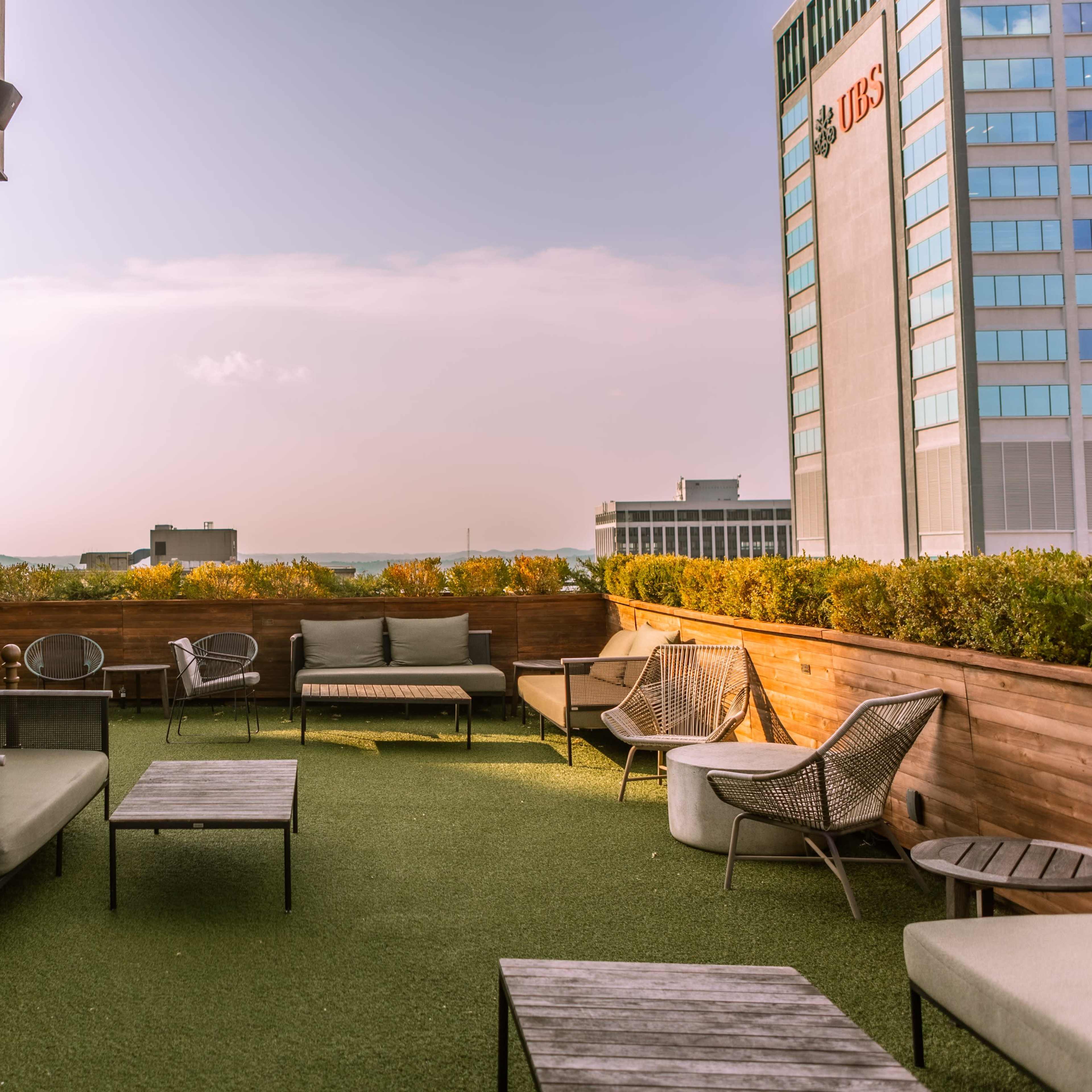 A rooftop lounge area with modern seating and greenery, set against a city skyline.