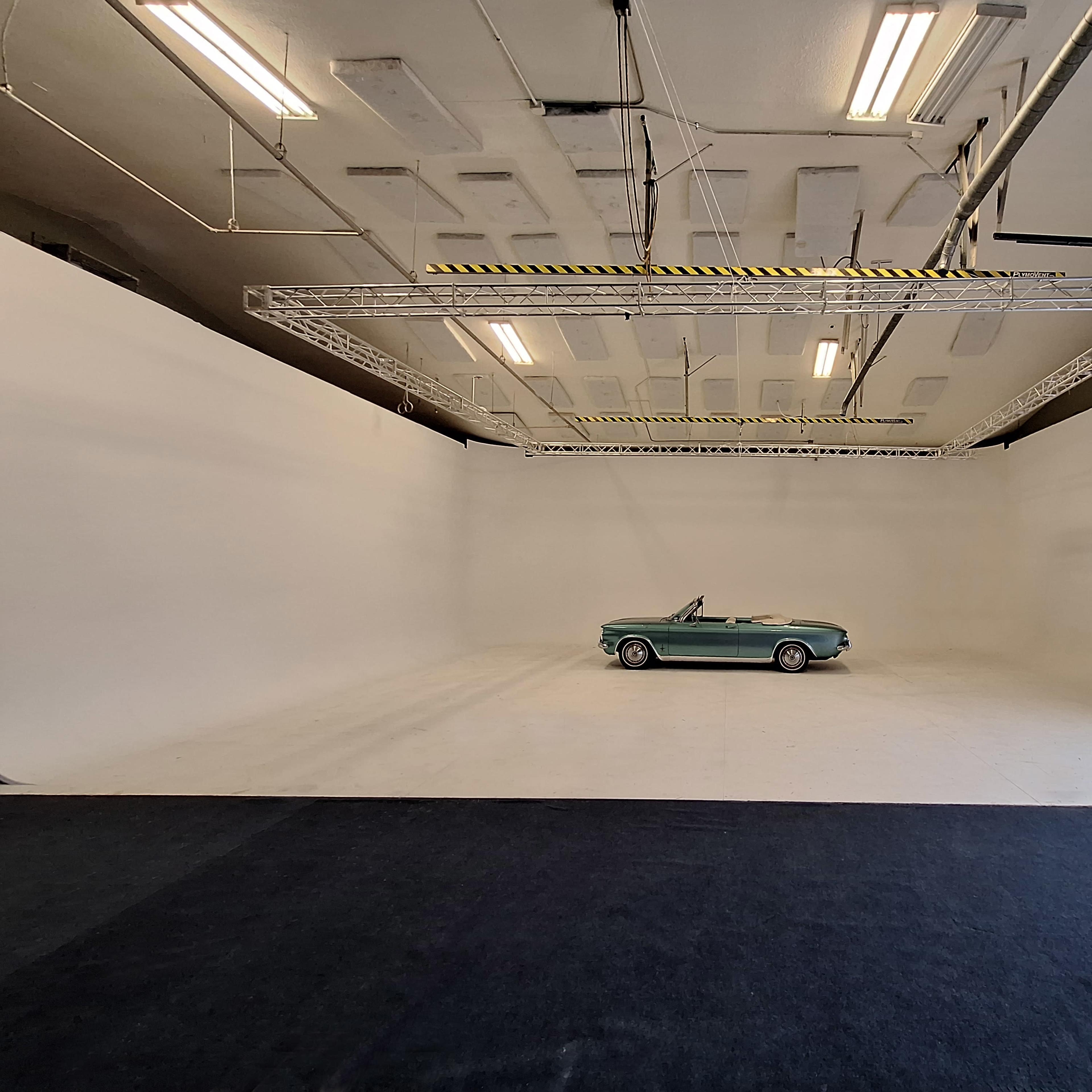 A vintage convertible car is positioned in a spacious, empty studio with white walls and overhead lighting.