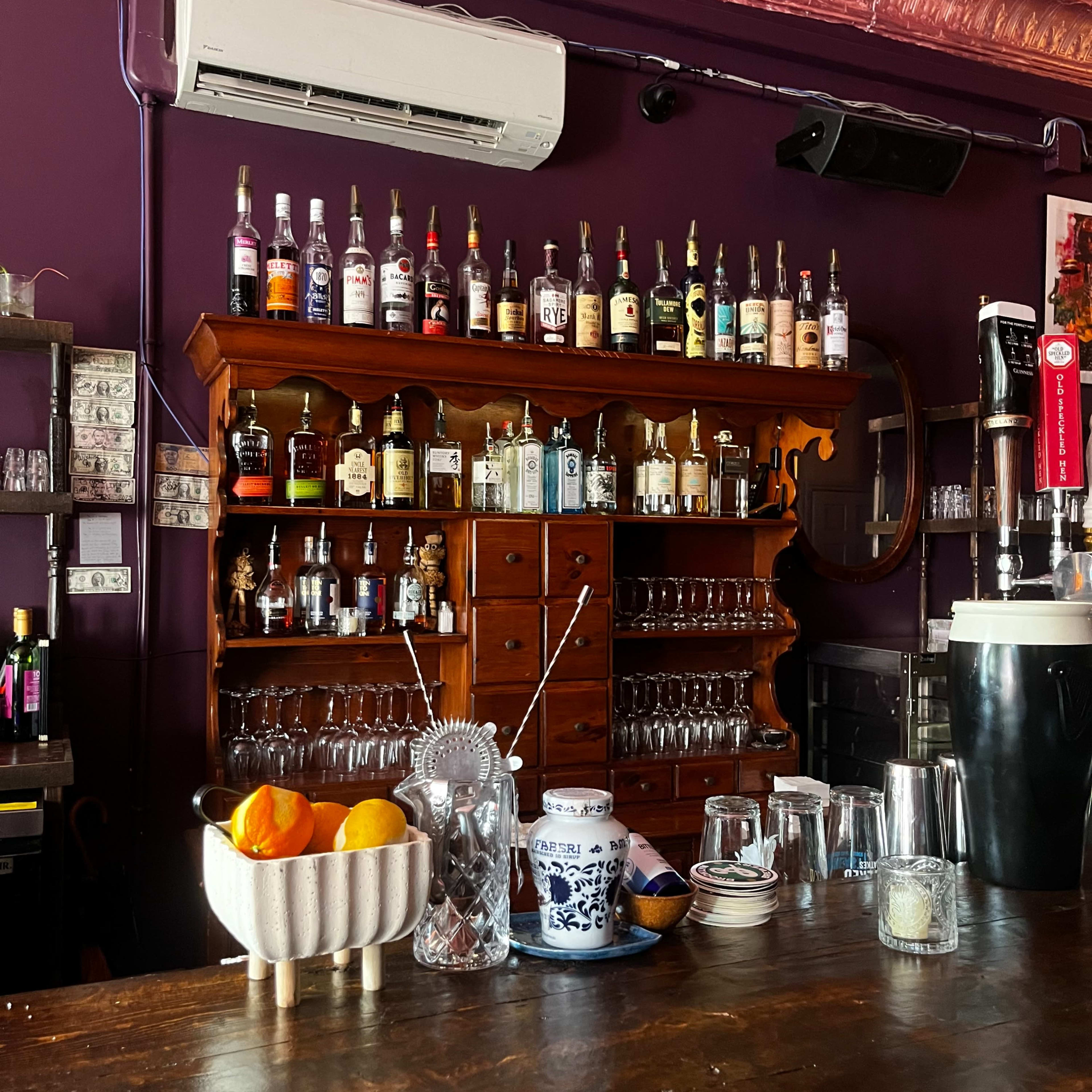 A bar with a wooden counter, an assortment of liquor bottles lined up on shelves above, and a bowl of oranges on the counter.