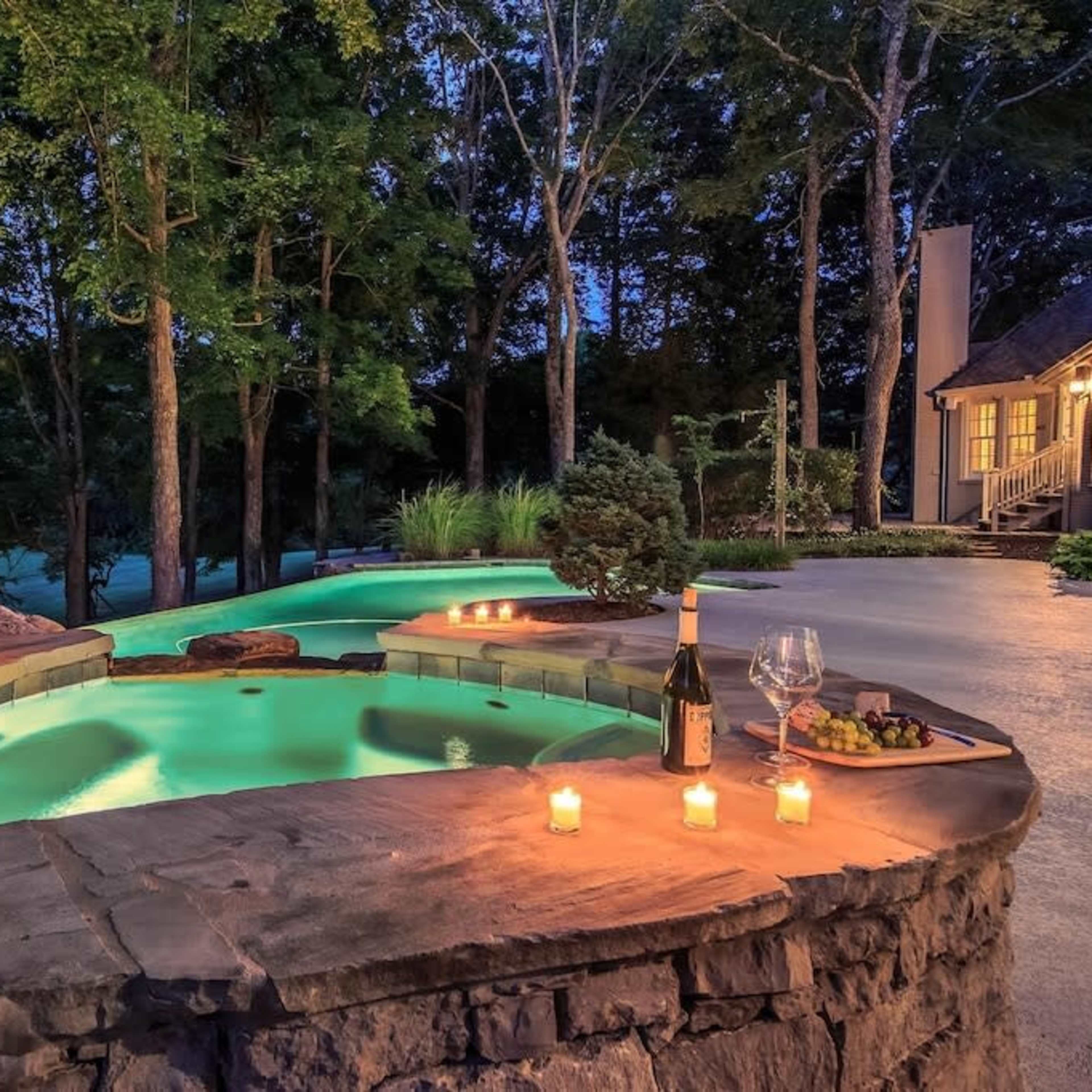 A stone patio features a circular hot tub with glowing water, surrounded by trees and softly lit candles, next to a bottle of wine and a glass on a small table.