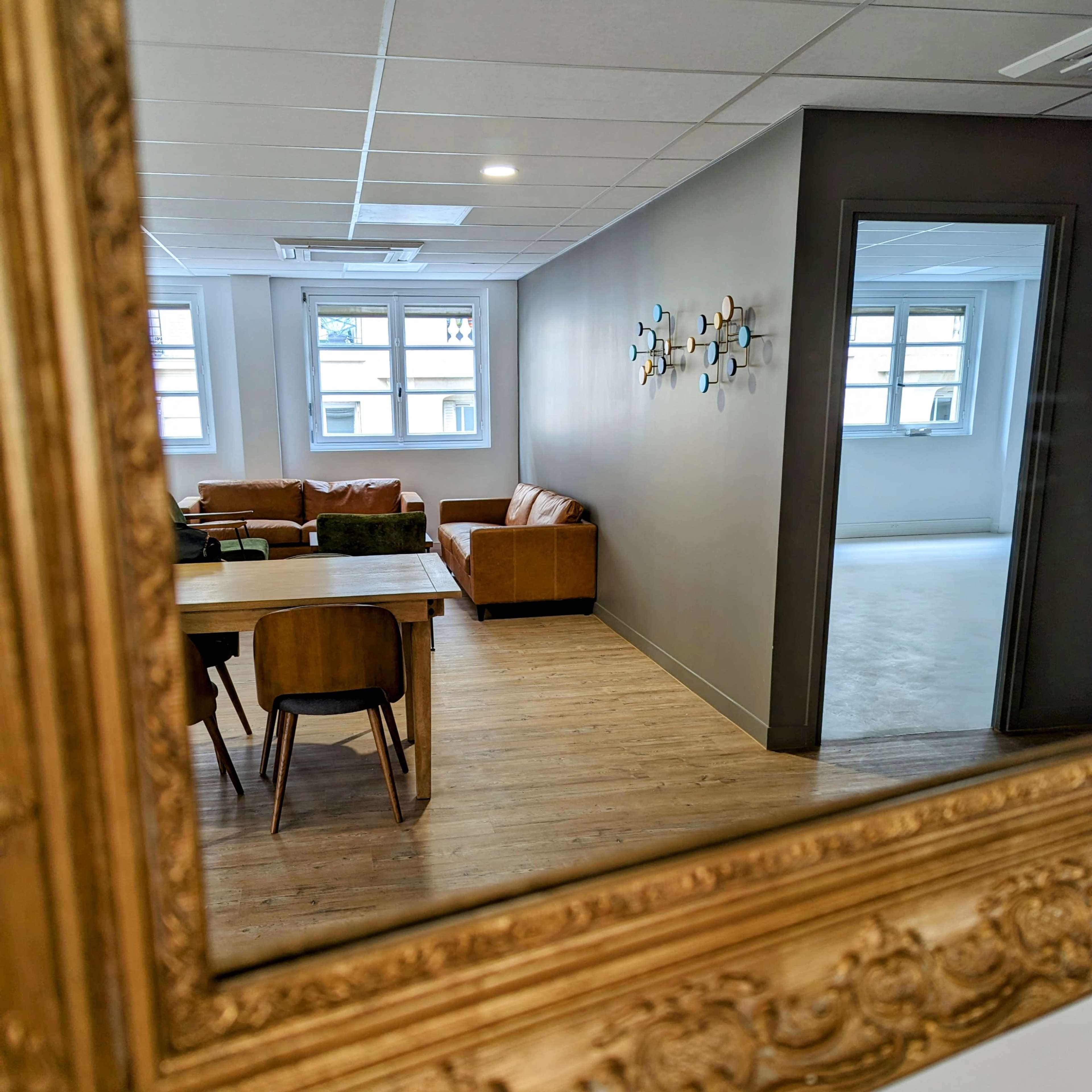 A elegantly framed mirror reflects a cozy lounge area with a brown sofa, wooden table, and chairs in a well-lit, minimalist room.
