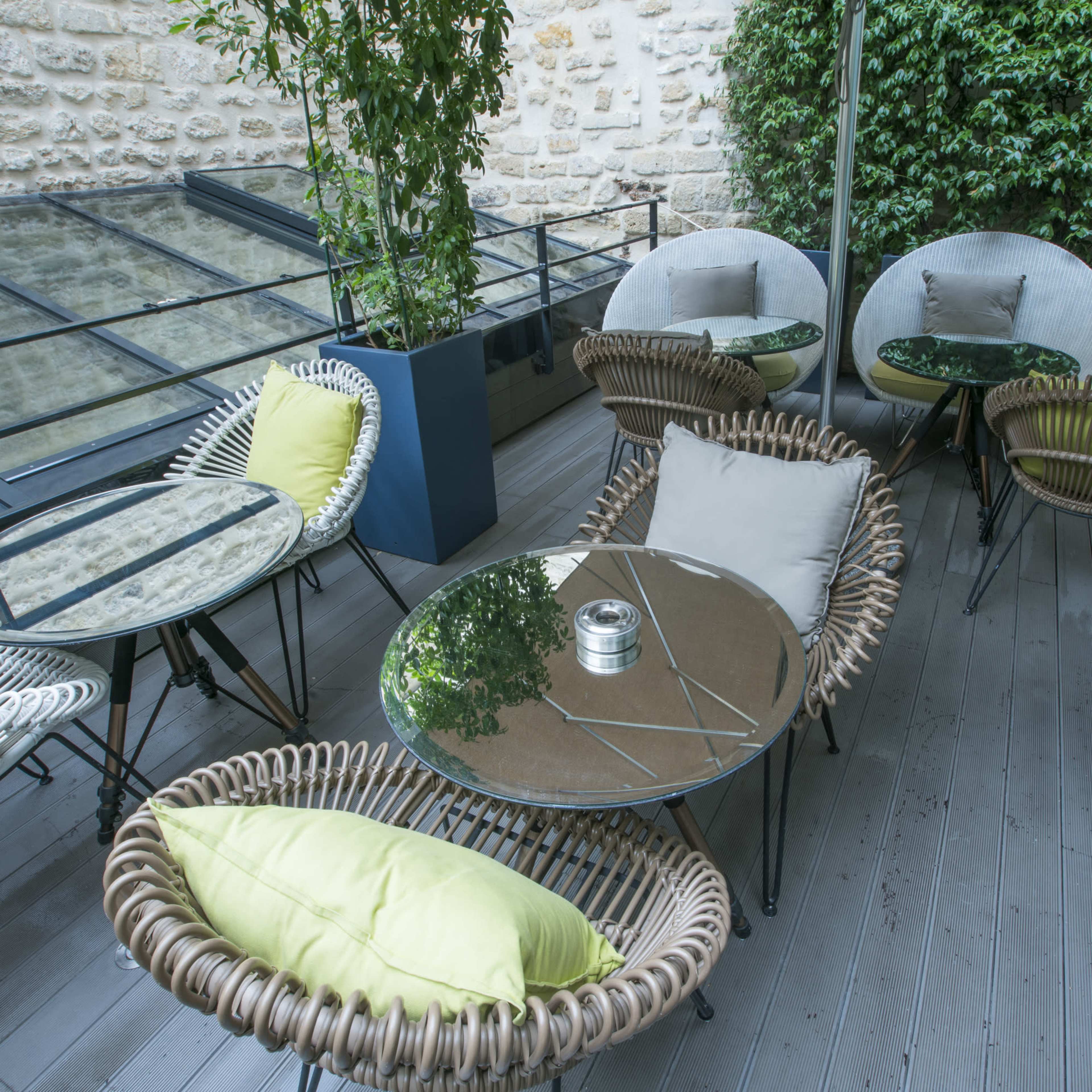 A patio area features round tables with glass tops surrounded by wicker chairs and green cushions, set against a stone wall.