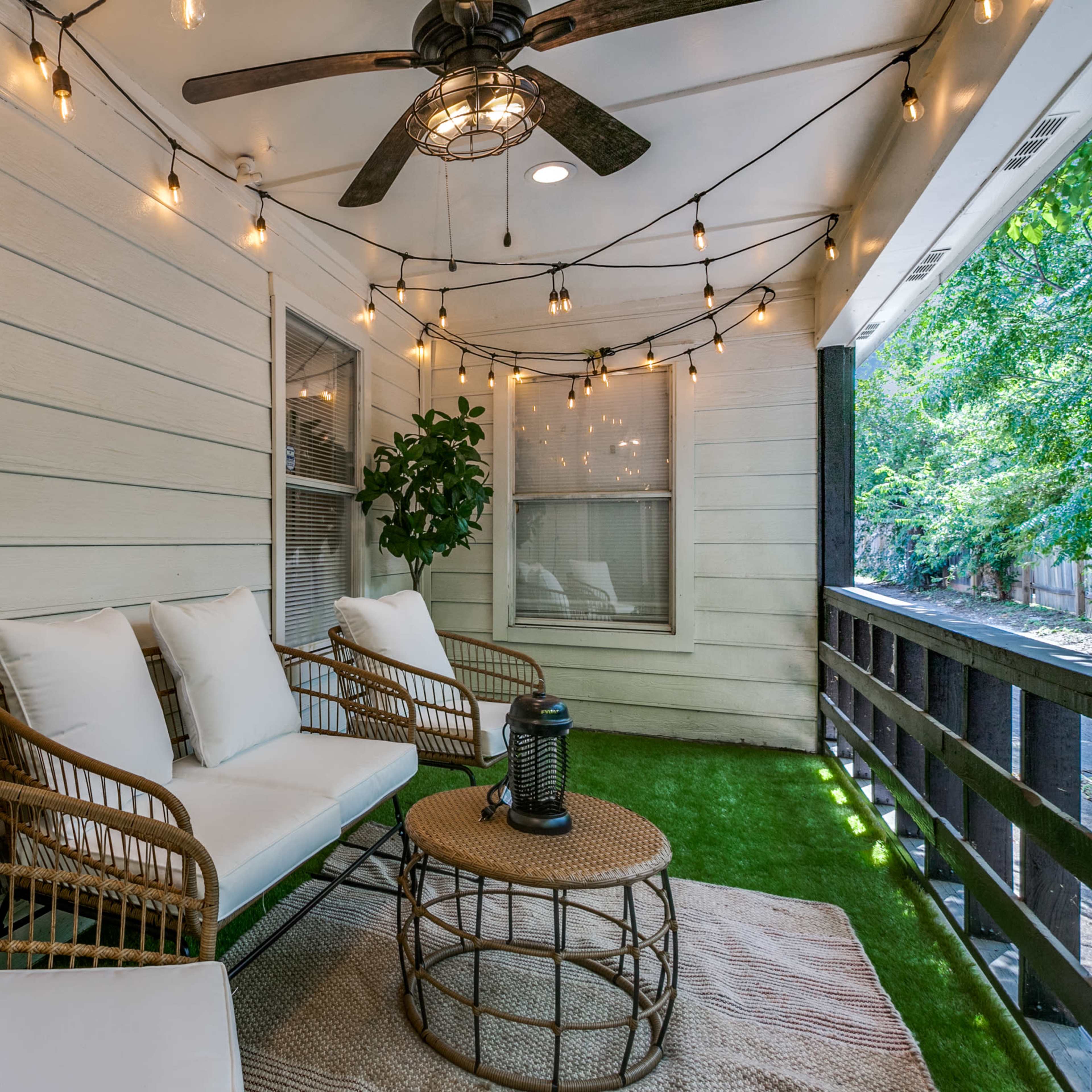 A cozy patio with white wicker furniture, a round table, and string lights hanging above, surrounded by greenery.