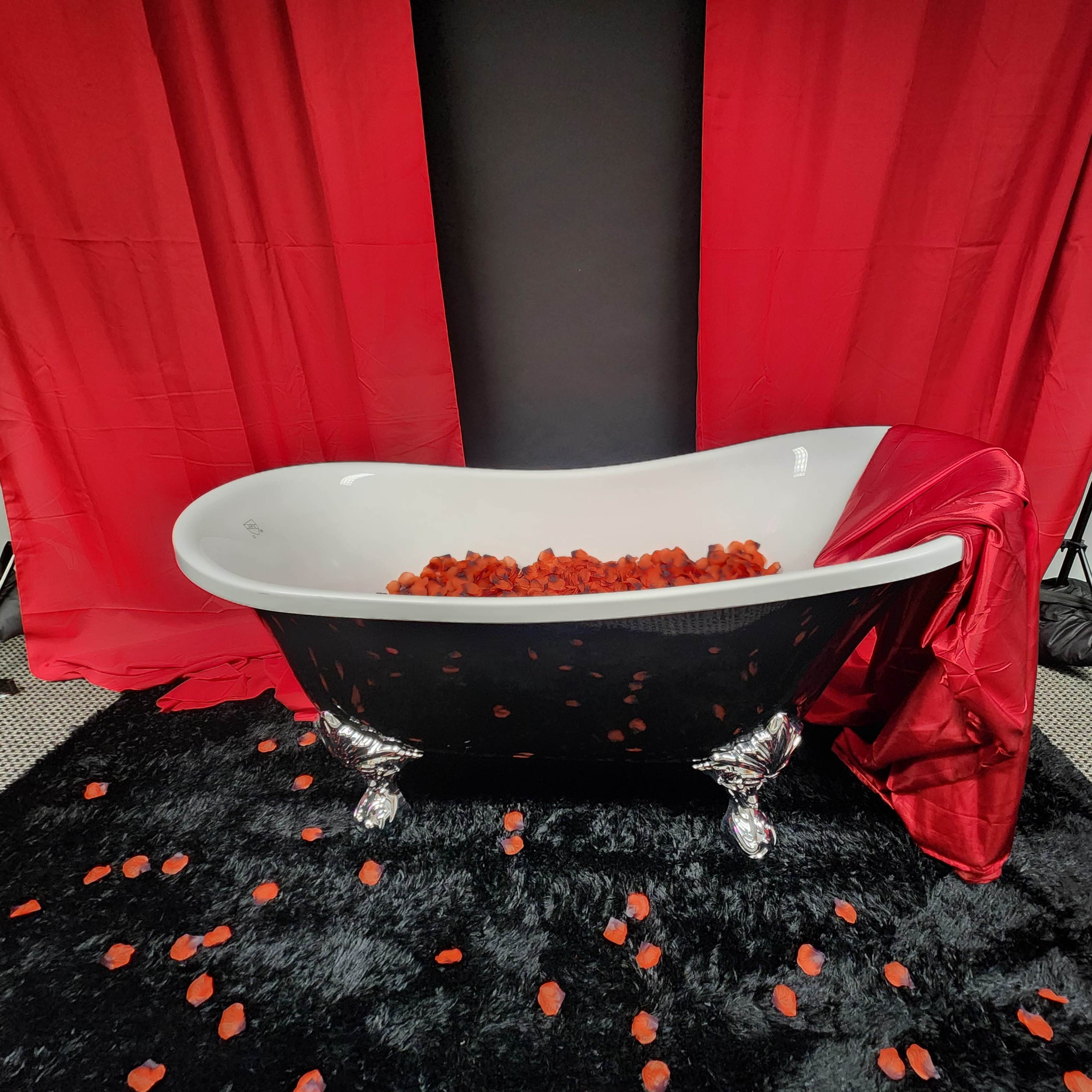 A black vintage bathtub filled with red rose petals sits on a plush black carpet, framed by red drapes in a studio setting.