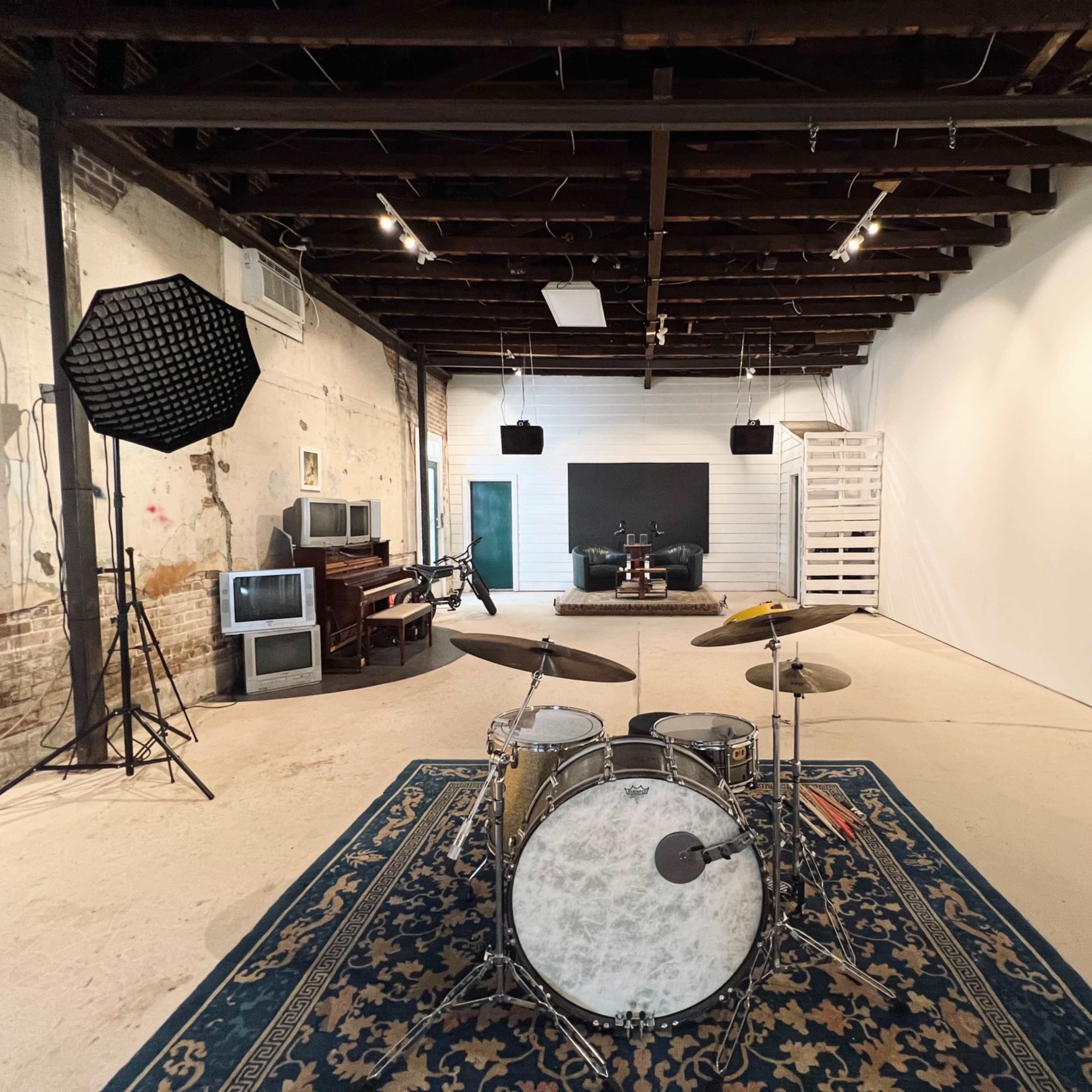 A spacious, minimalist studio with a drum set in the foreground, surrounded by various audio equipment and a blank wall in the background.