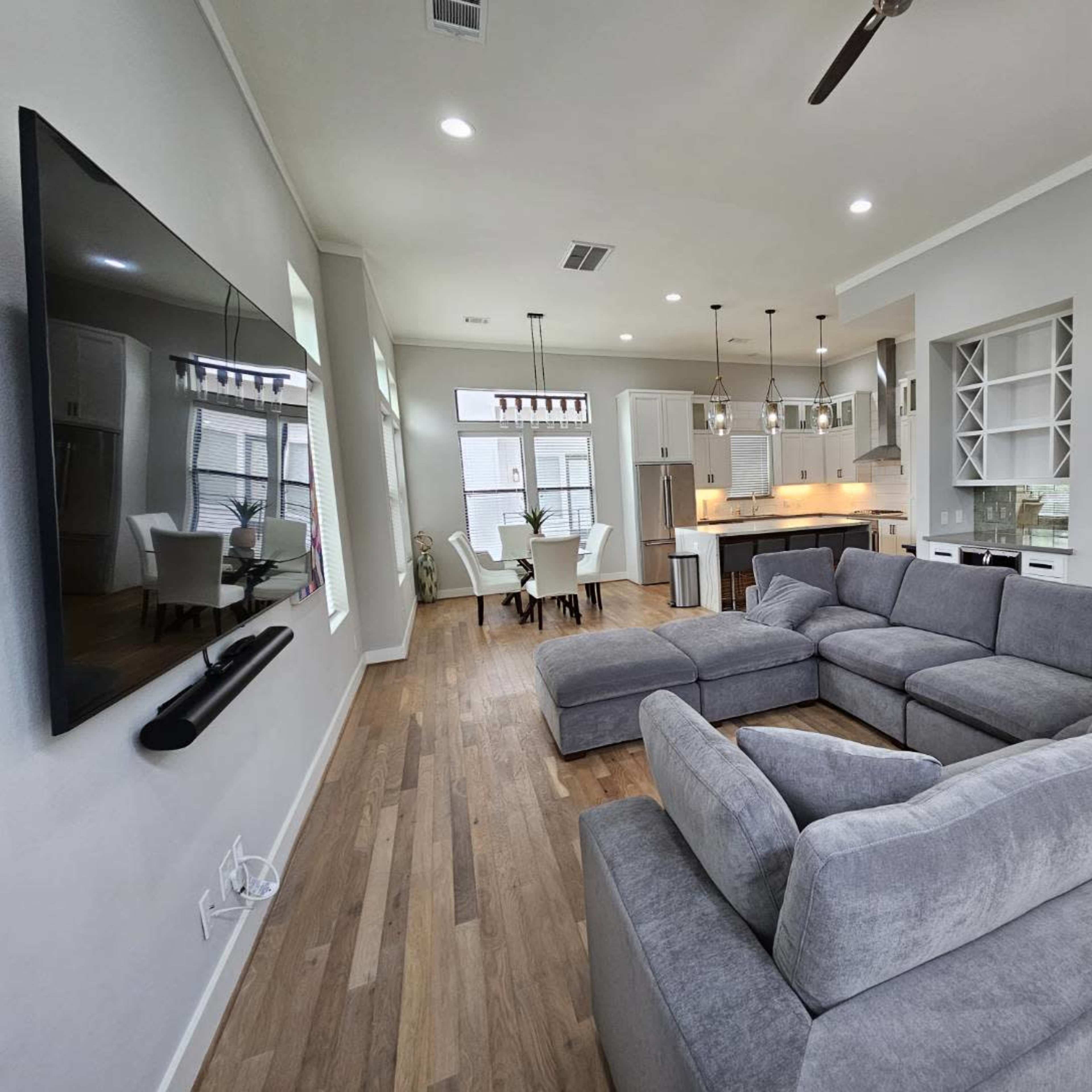 A modern living room with a large sectional sofa facing a wall-mounted television, leading into an open kitchen area with a dining table.