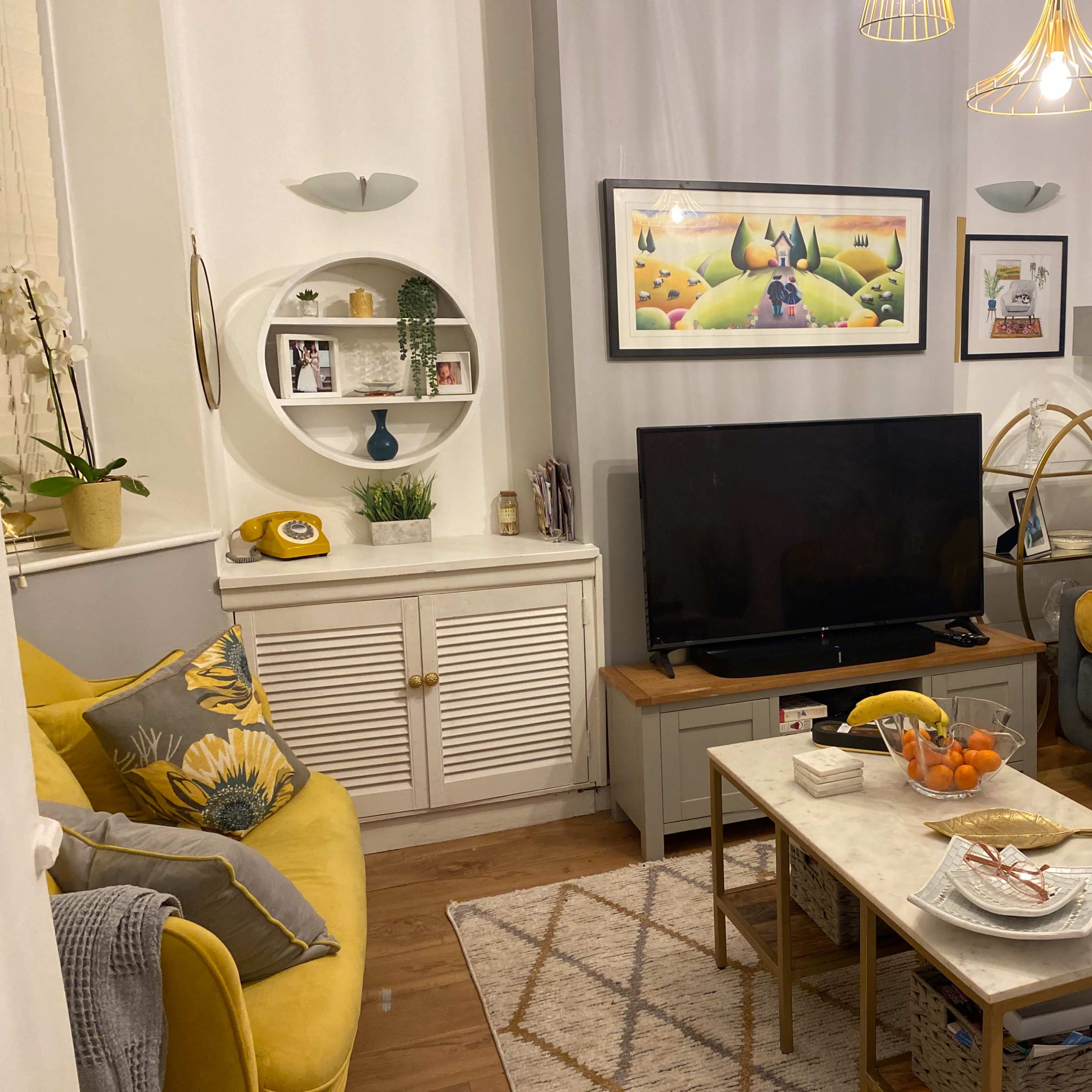 A cozy living room with a gray sofa, a yellow chair, a television on a wooden stand, and a coffee table with snacks, surrounded by decorative elements like plants and artwork.