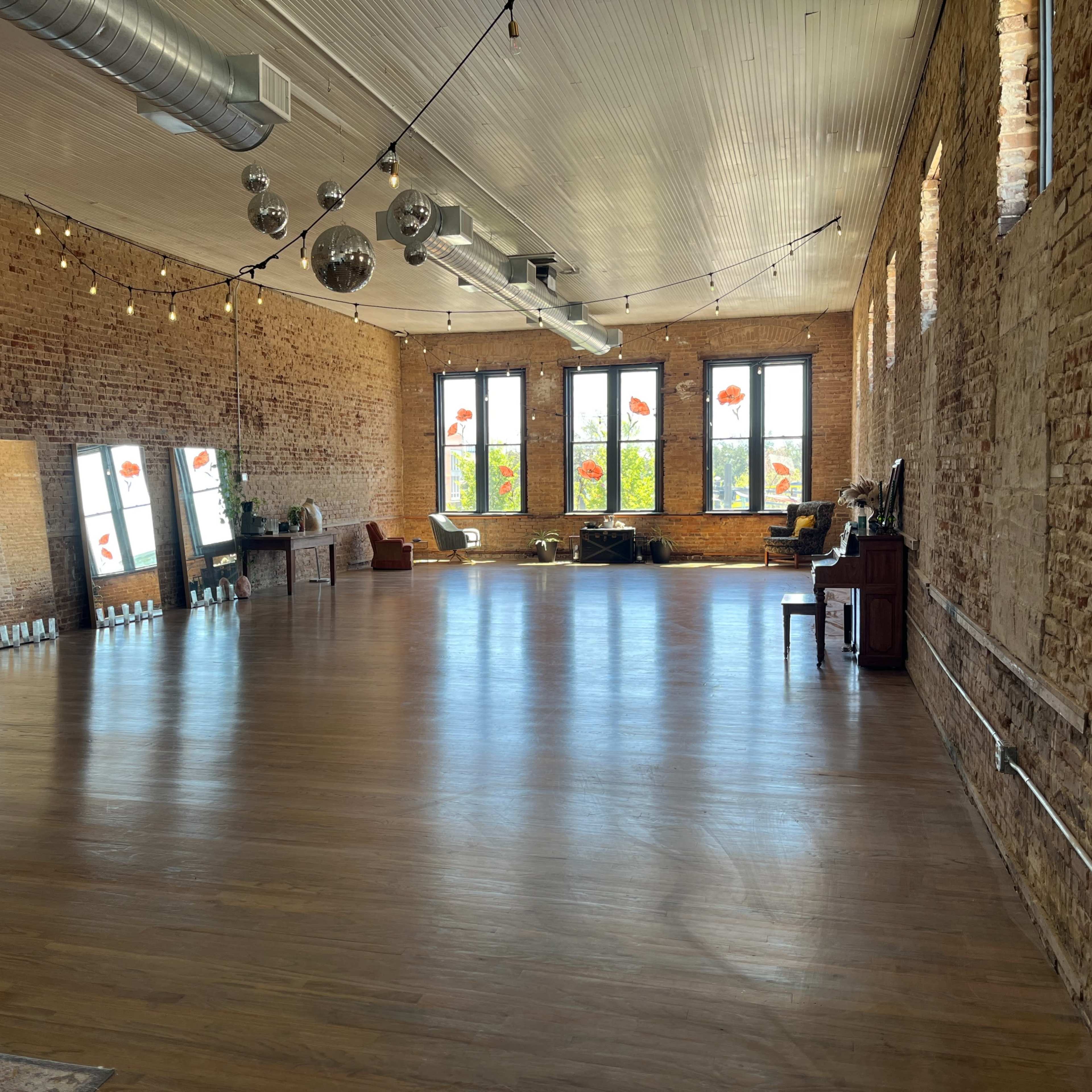 A spacious room with exposed brick walls, large windows, and wooden flooring, featuring mirrors along one side and a piano in the corner.