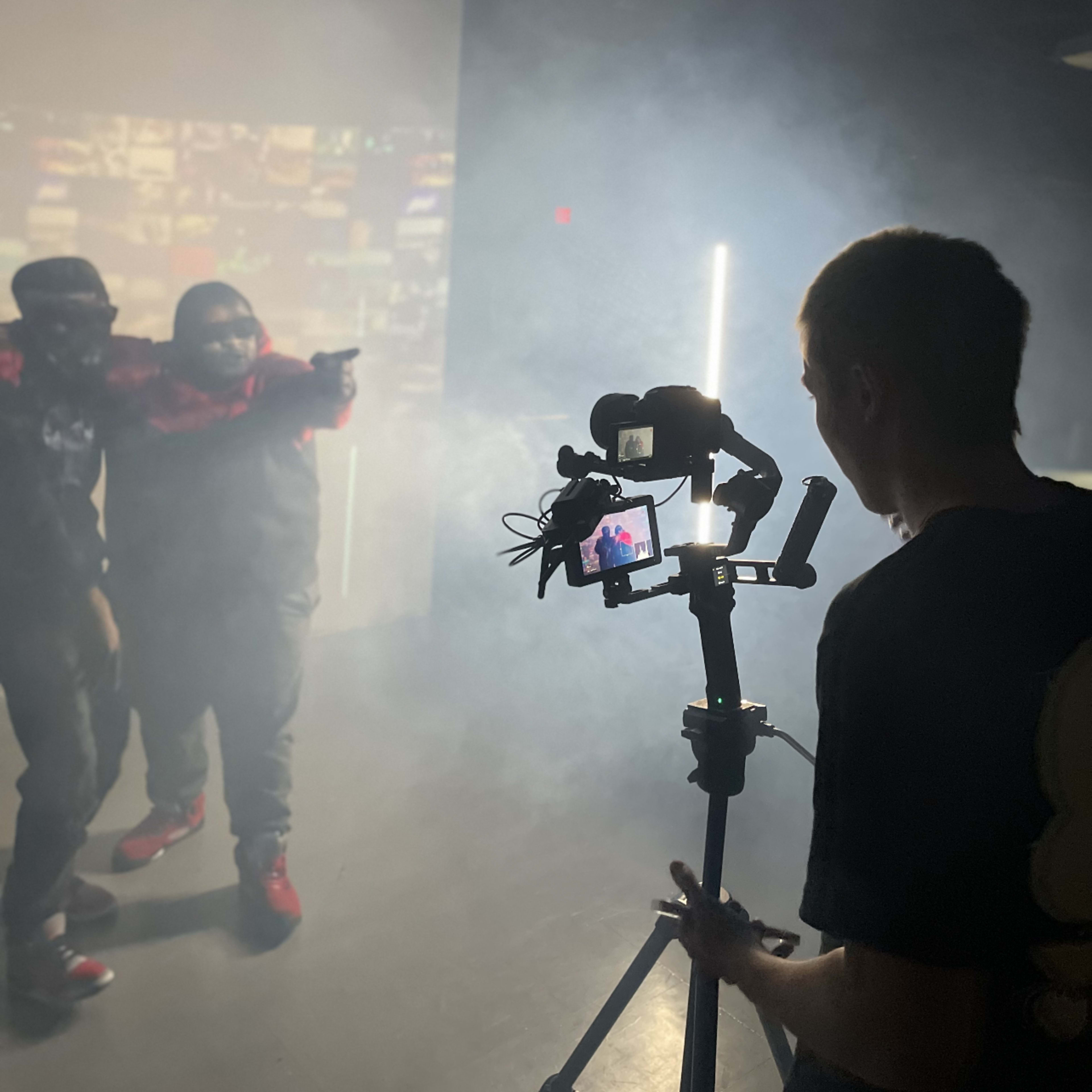 A person operates a camera on a gimbal while two individuals pose in a smoky setting with a projected background.