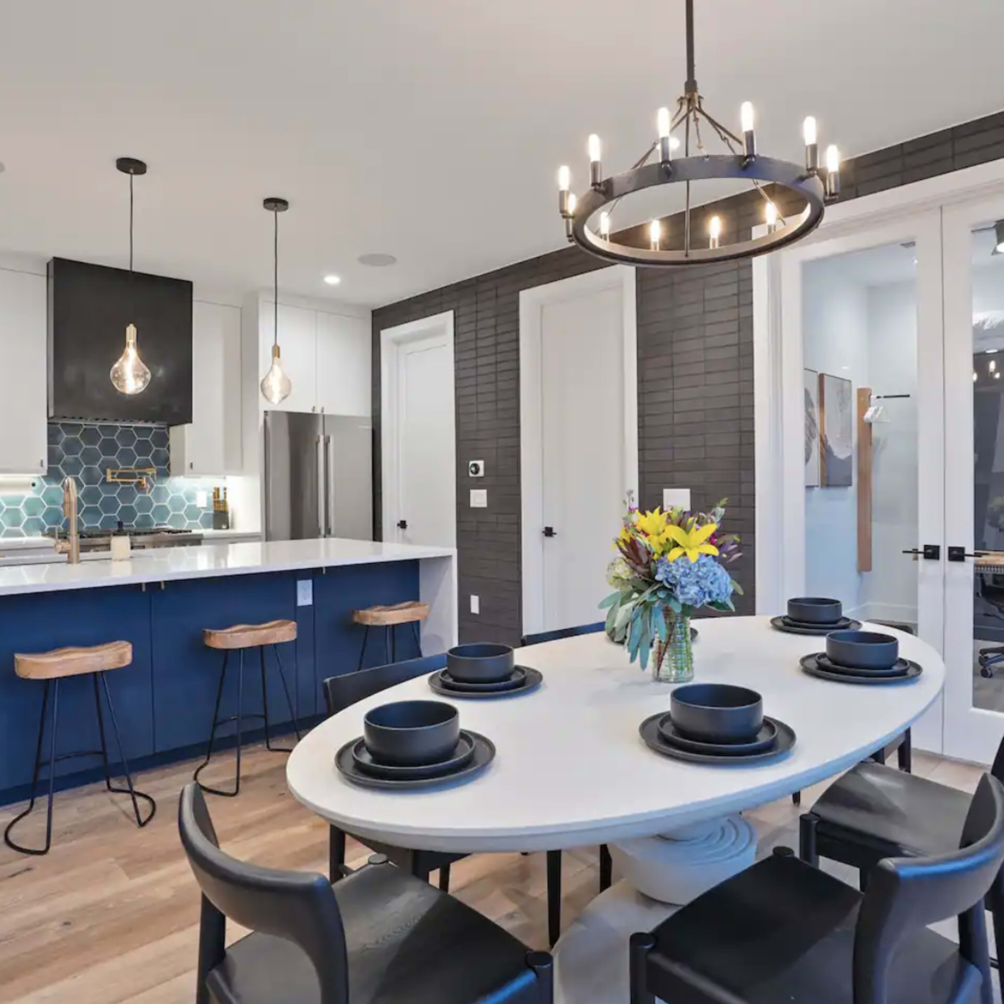 A modern kitchen features a round dining table set for six, with bar stools at the countertop and pendant lights above the island.