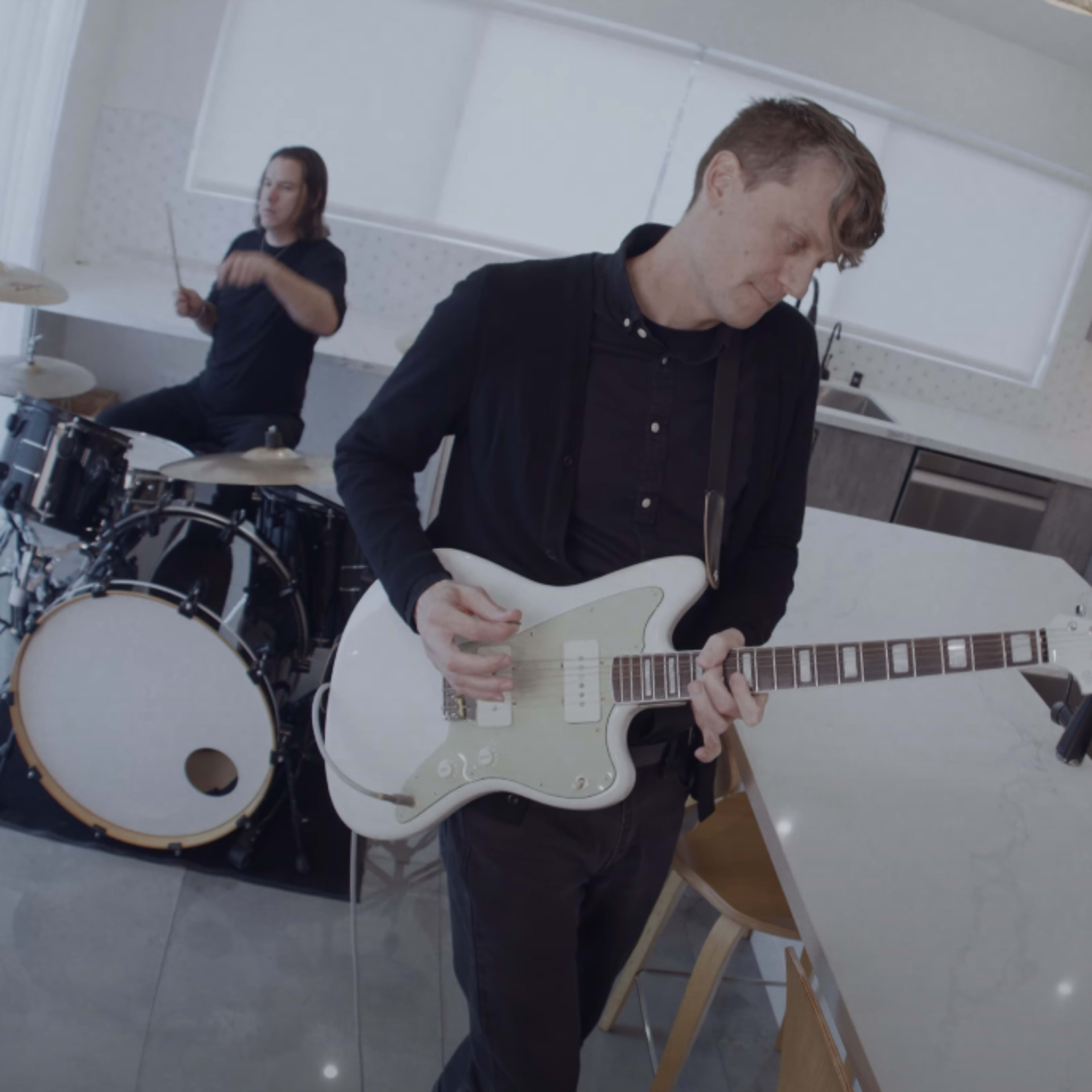A band performs in a modern kitchen with a drummer, guitarist, and bassist.