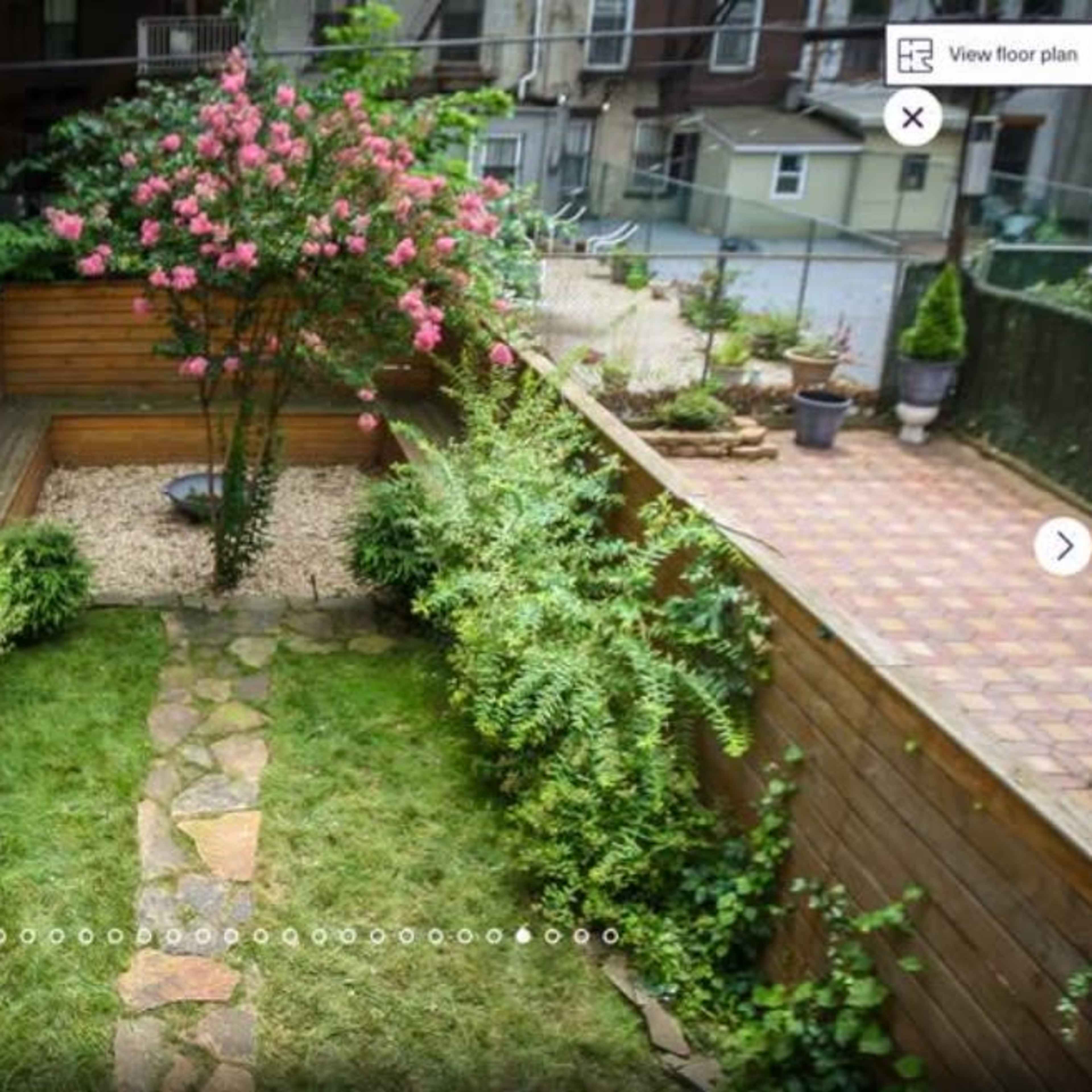 A landscaped garden featuring a stone pathway leading to a flowering shrub, surrounded by various plants and gravel areas.