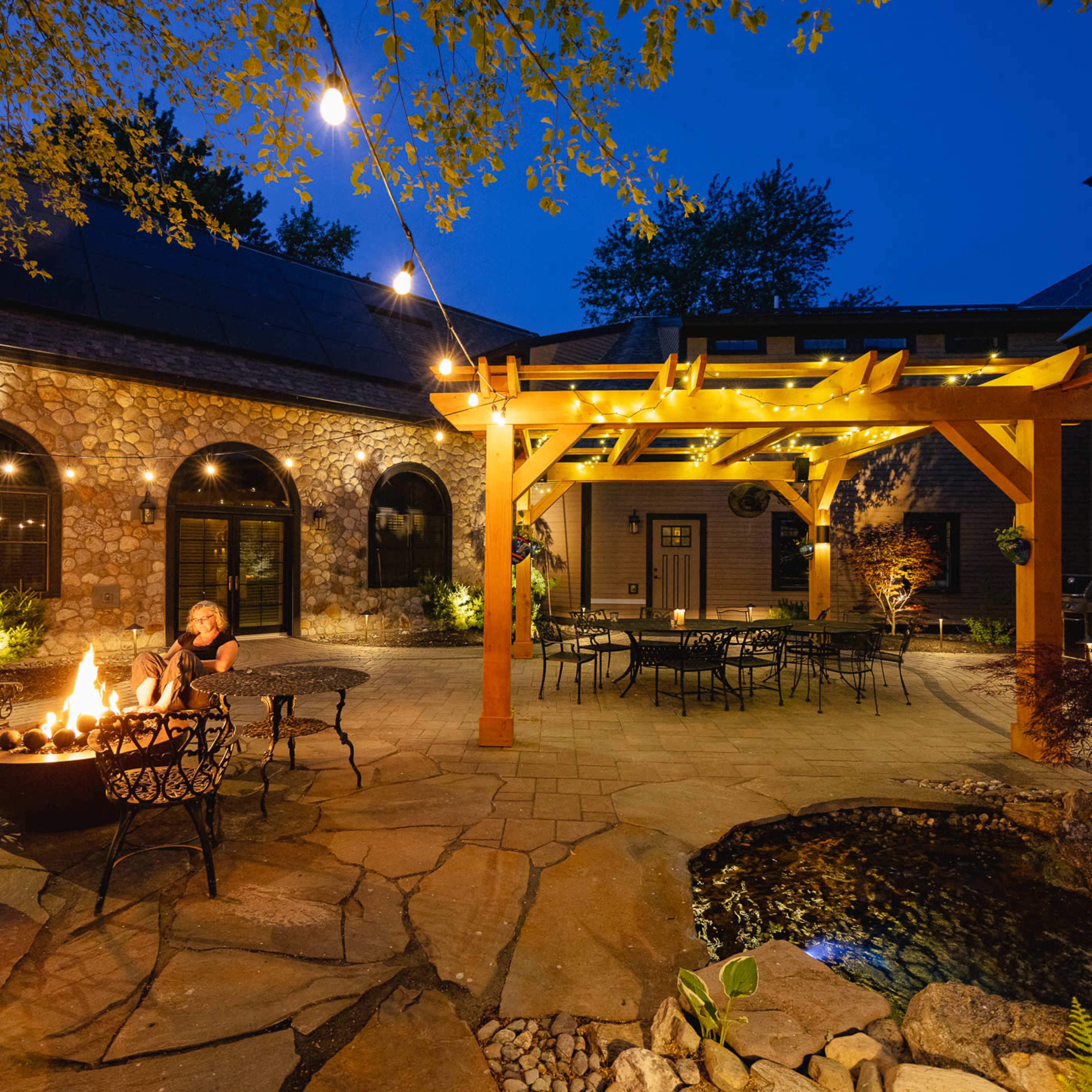 A stone patio features a fire pit surrounded by chairs, with a pergola illuminated by string lights and a small pond nearby, all set in the evening.