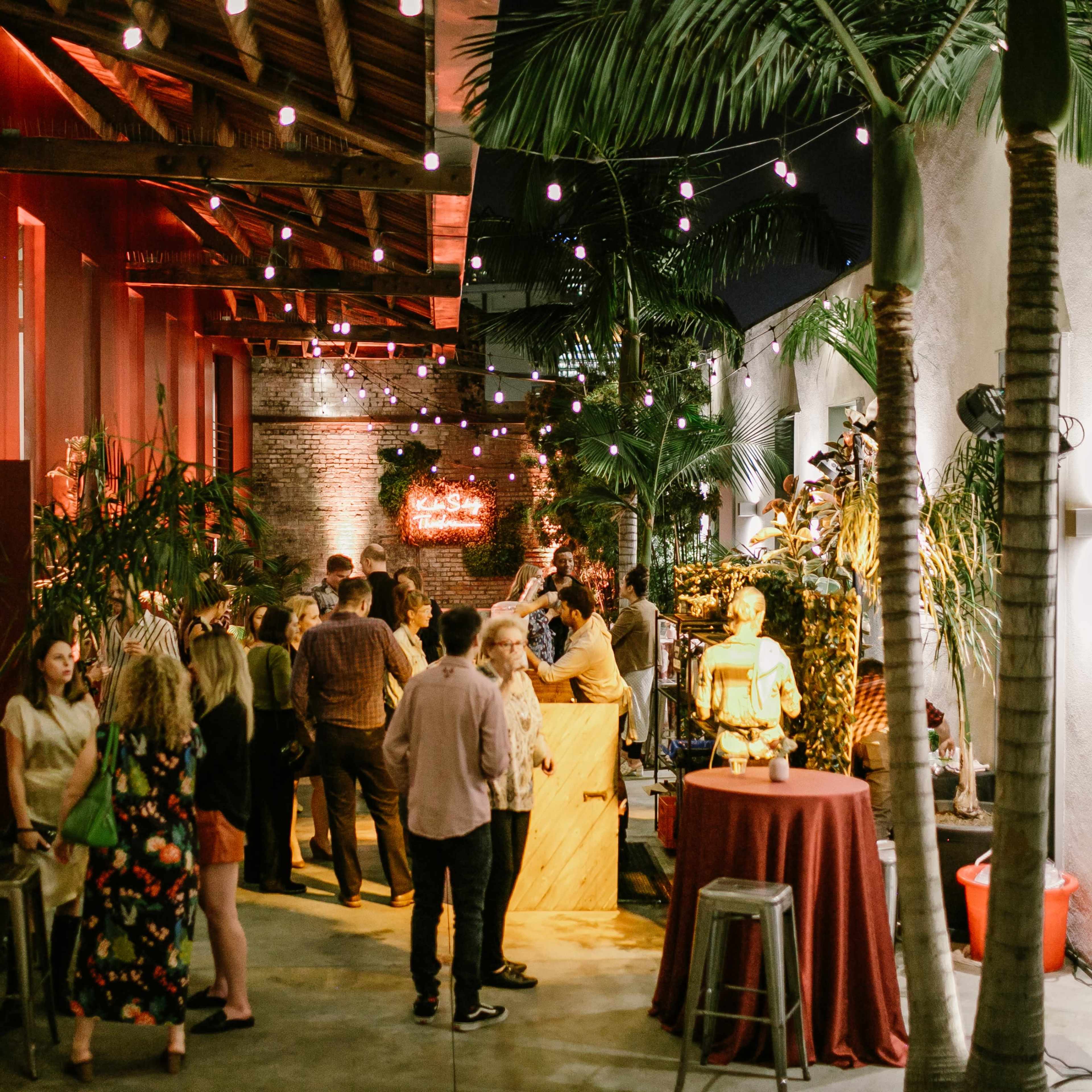 A group of people socializes at a lively indoor event space decorated with plants and string lights.