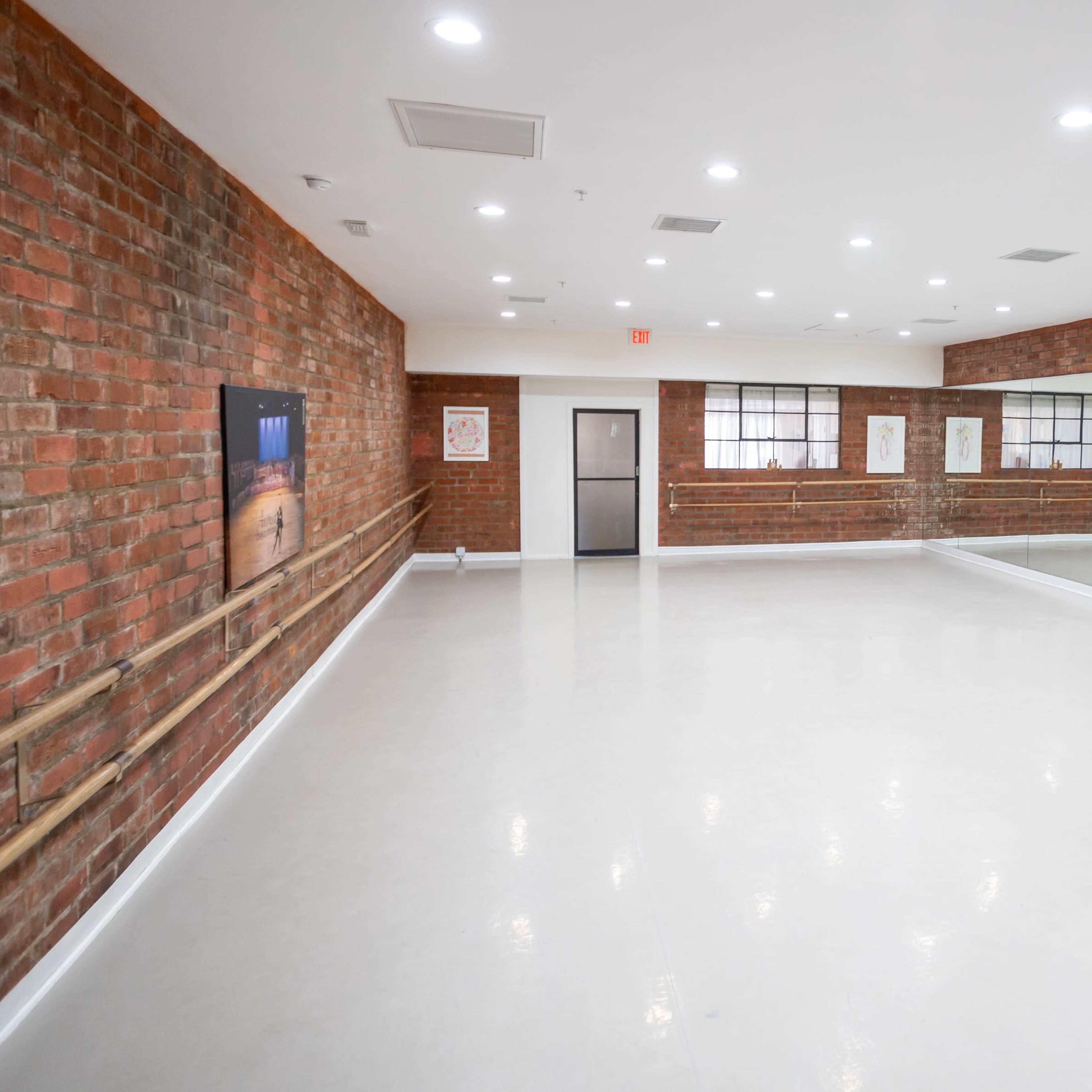 An empty dance studio with white floors, exposed brick walls, and mirrors on one side.