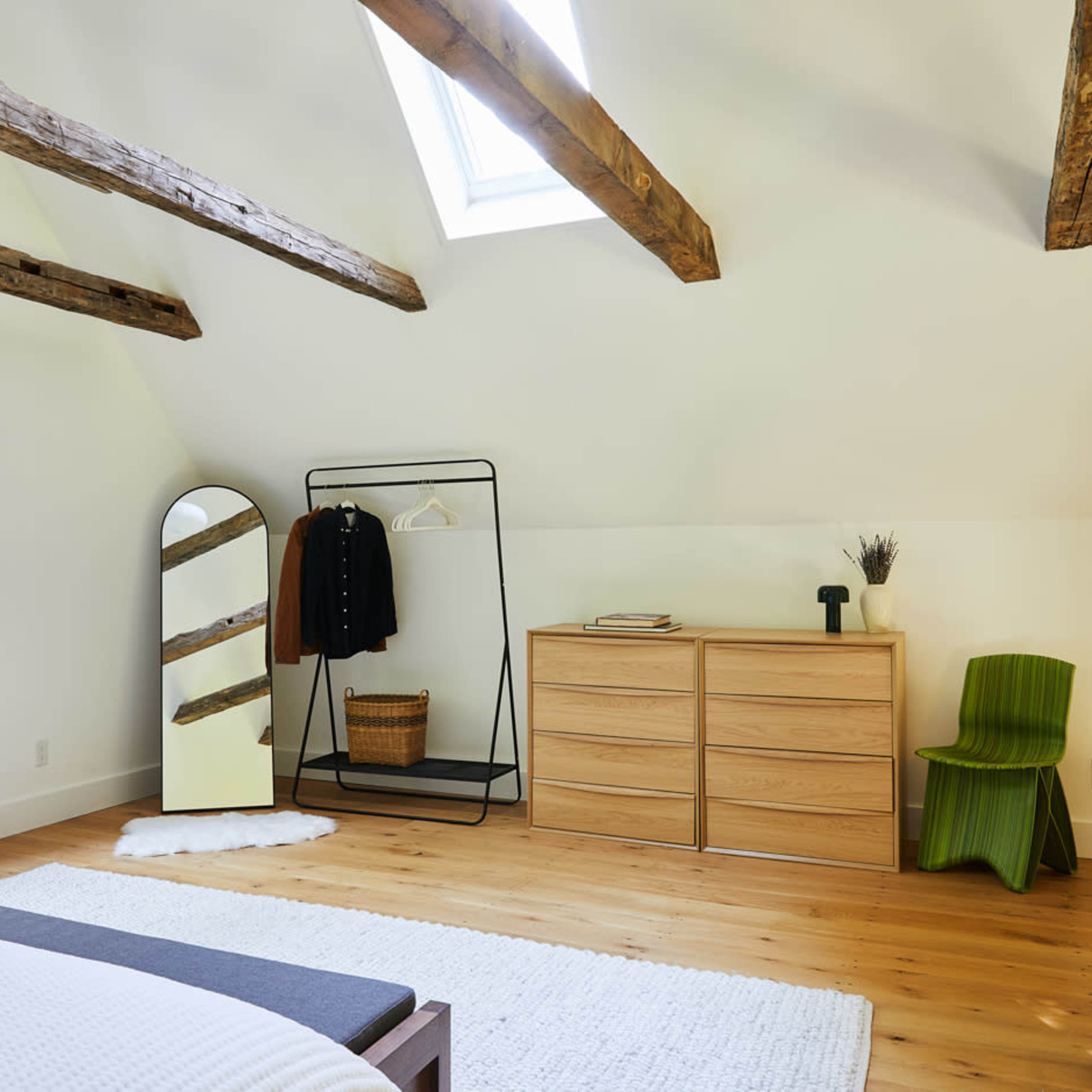 The bedroom features a sloped ceiling with exposed wooden beams, a large window, a minimalist wardrobe, a wooden chest of drawers, and a distinct green chair.