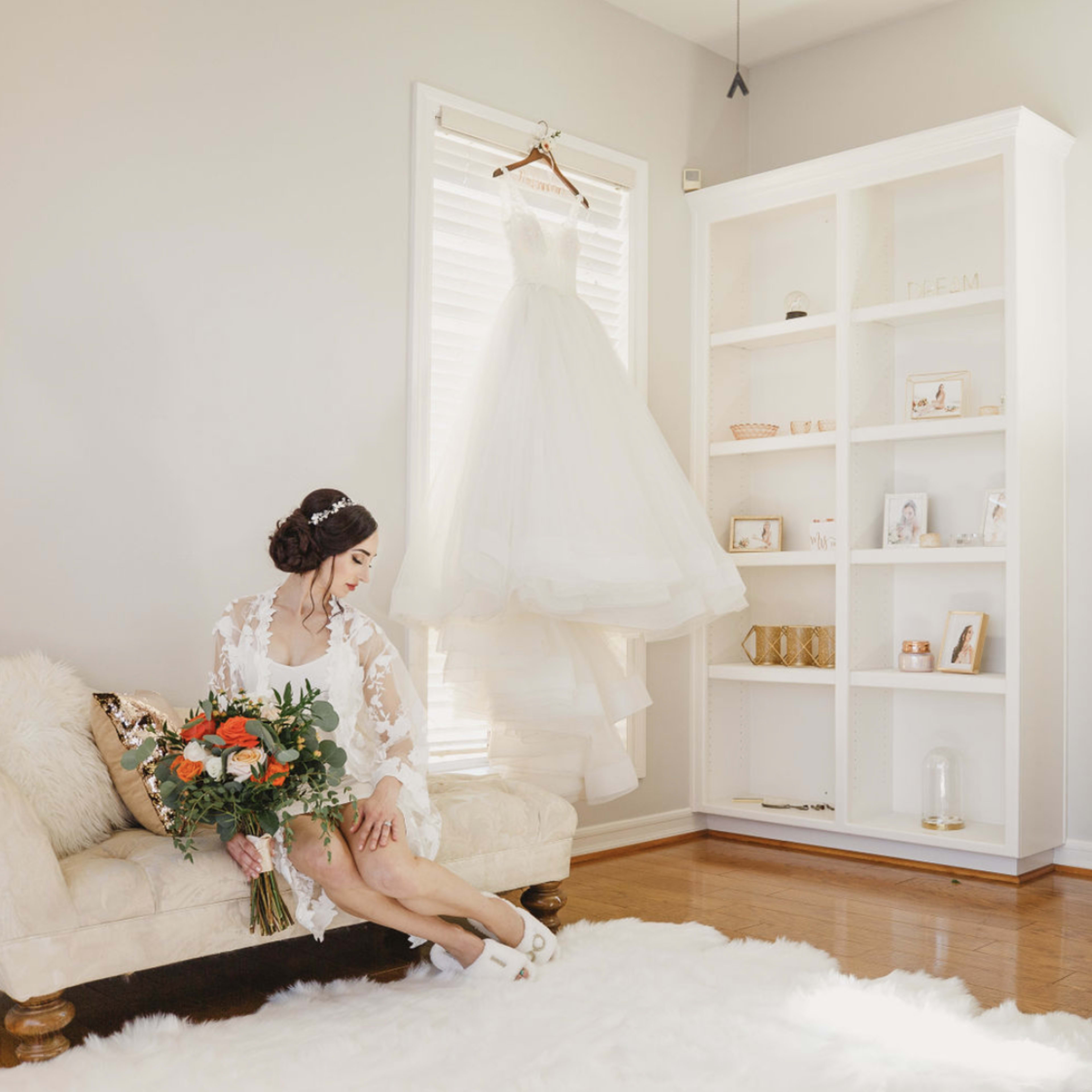 A bride in a white dress sits on a couch holding a bouquet of flowers, while her wedding gown hangs on a rack in a well-lit room.