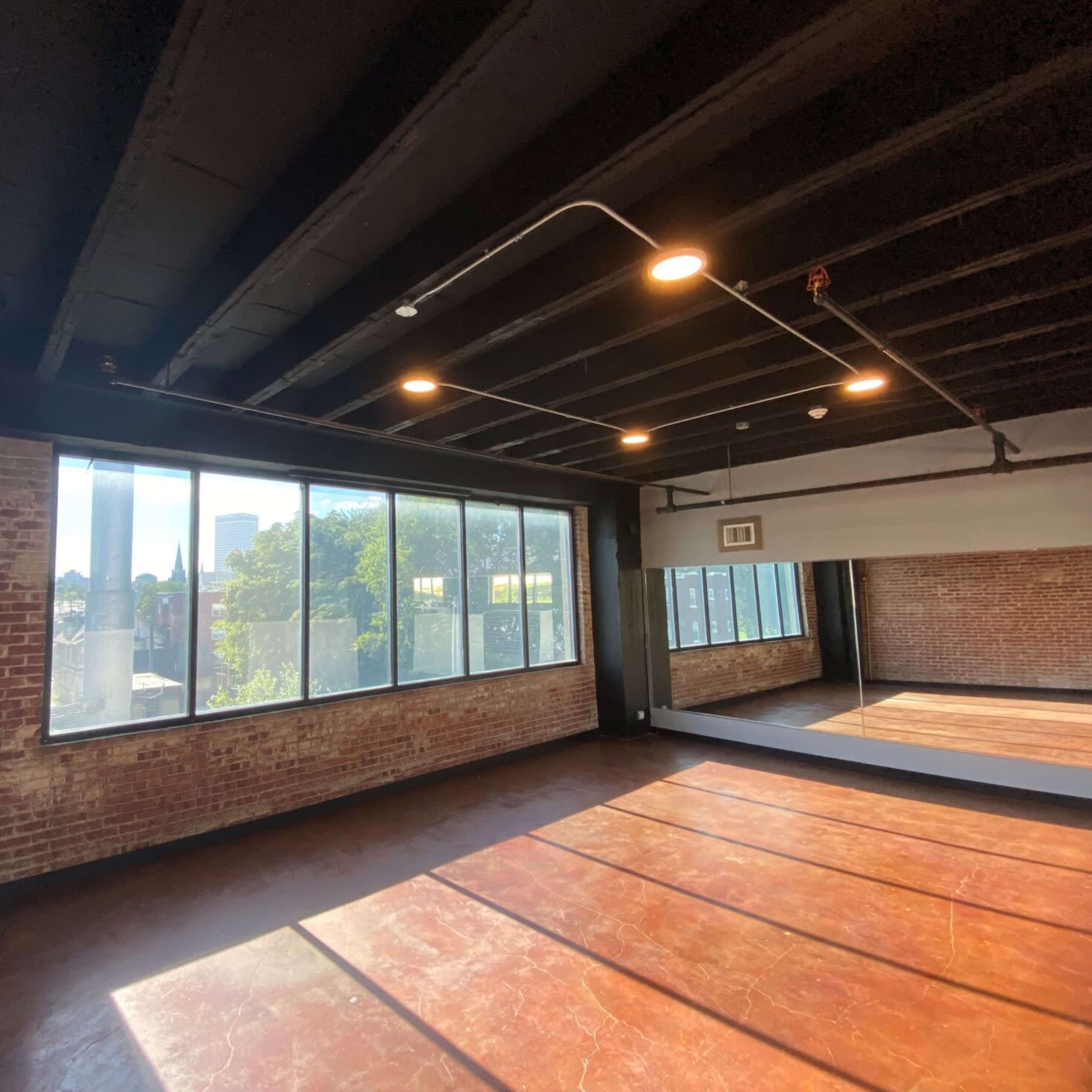 A spacious room with exposed brick walls, large windows, and a hardwood floor, featuring overhead lighting and a mirror on one side.