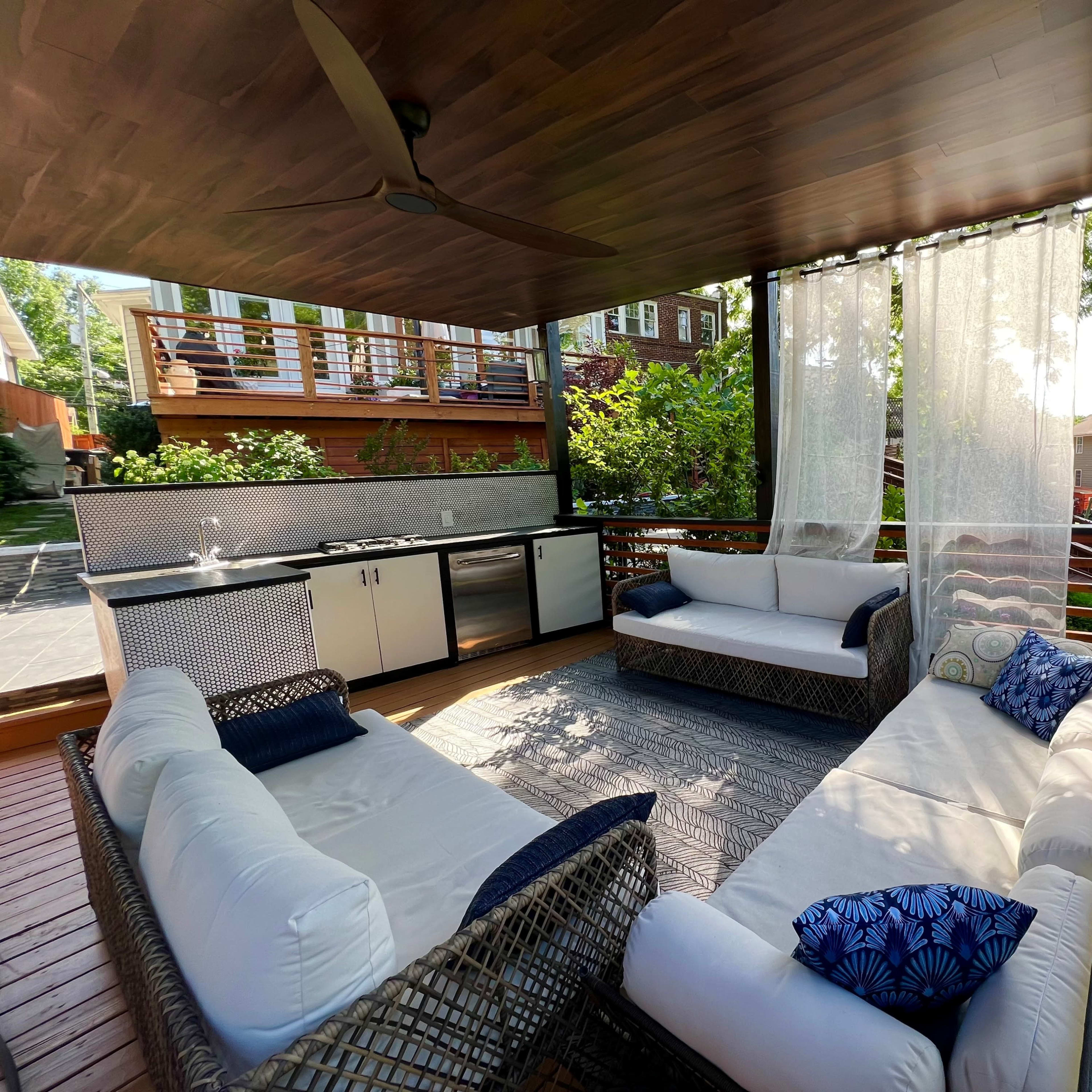 A covered outdoor seating area with a kitchen and comfortable sofas surrounded by greenery.