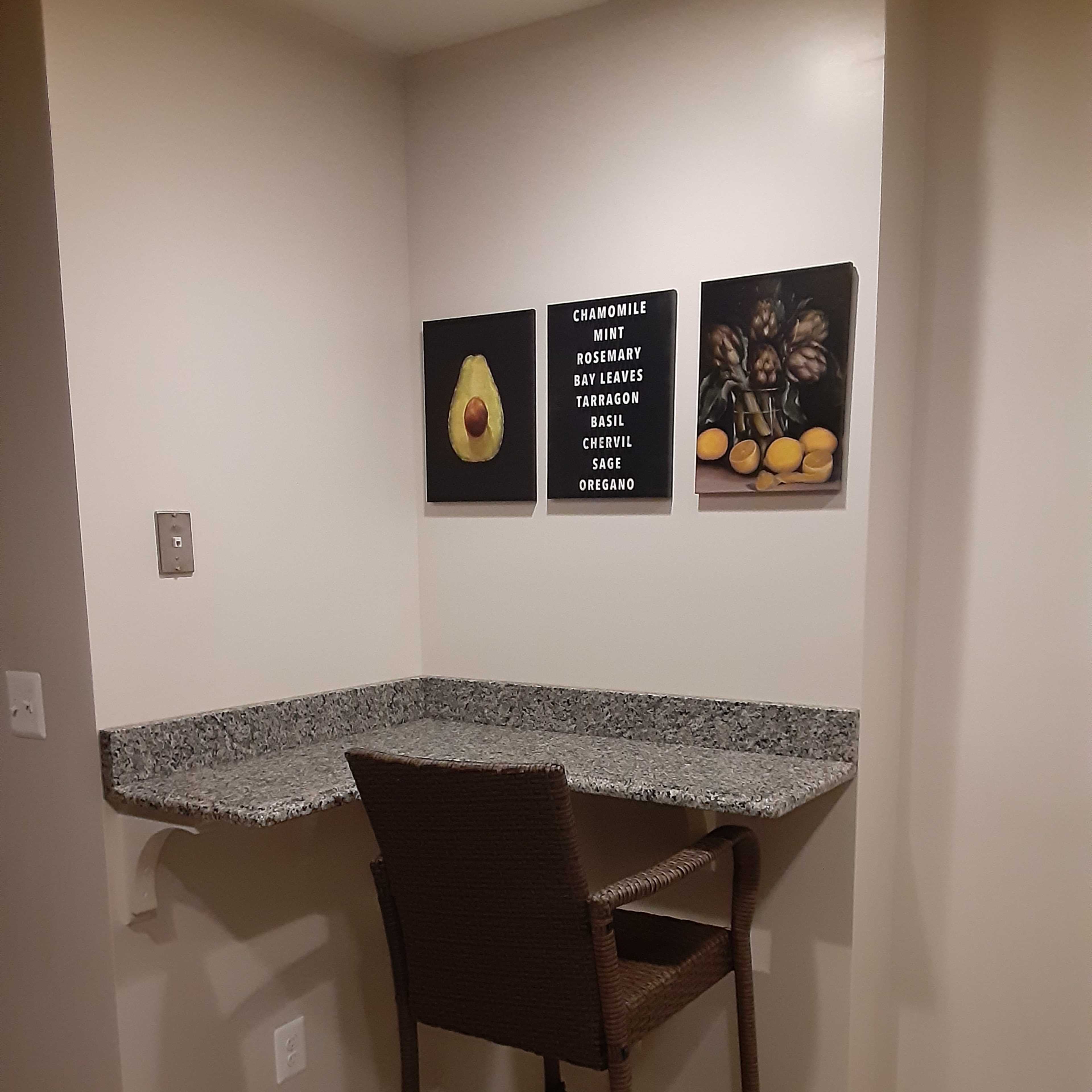 A corner nook features a granite countertop and a single barstool, with three framed images of vegetables and herbs hanging on the wall above.