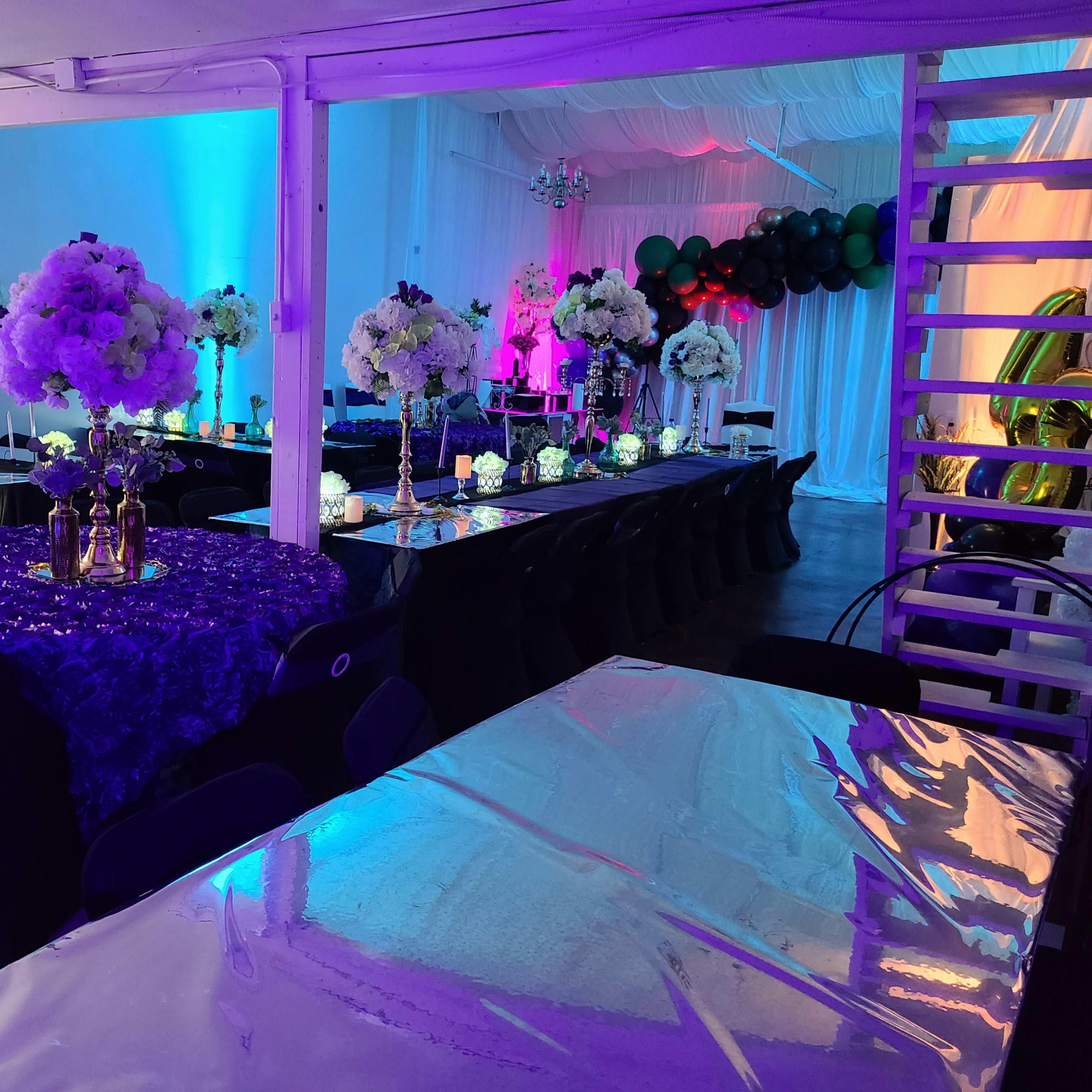 A decorated event space with tables set for a gathering, featuring floral arrangements, colorful lighting, and a balloon arch.