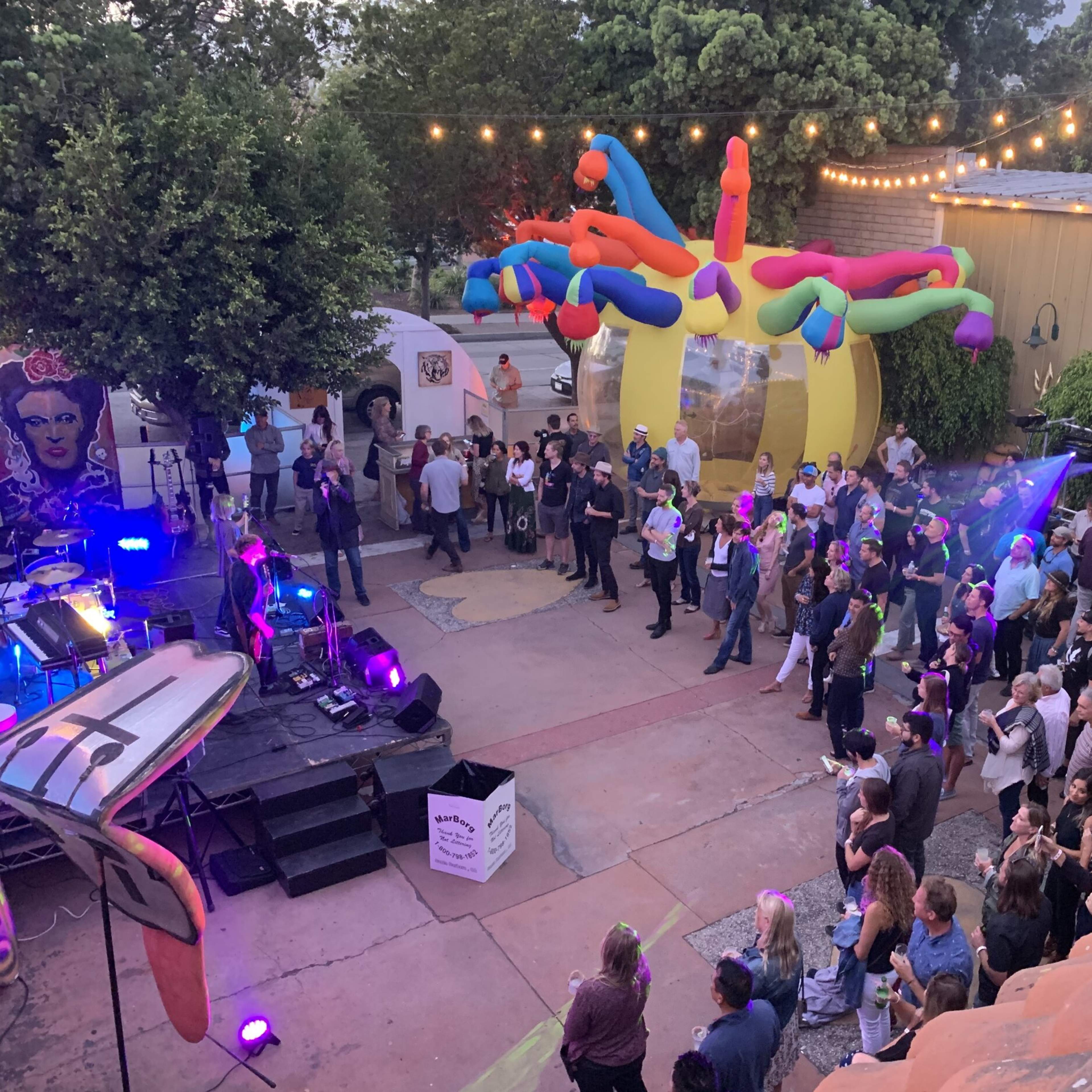 A lively outdoor gathering features a crowd enjoying a performance near colorful inflatable decorations and string lights hanging overhead.