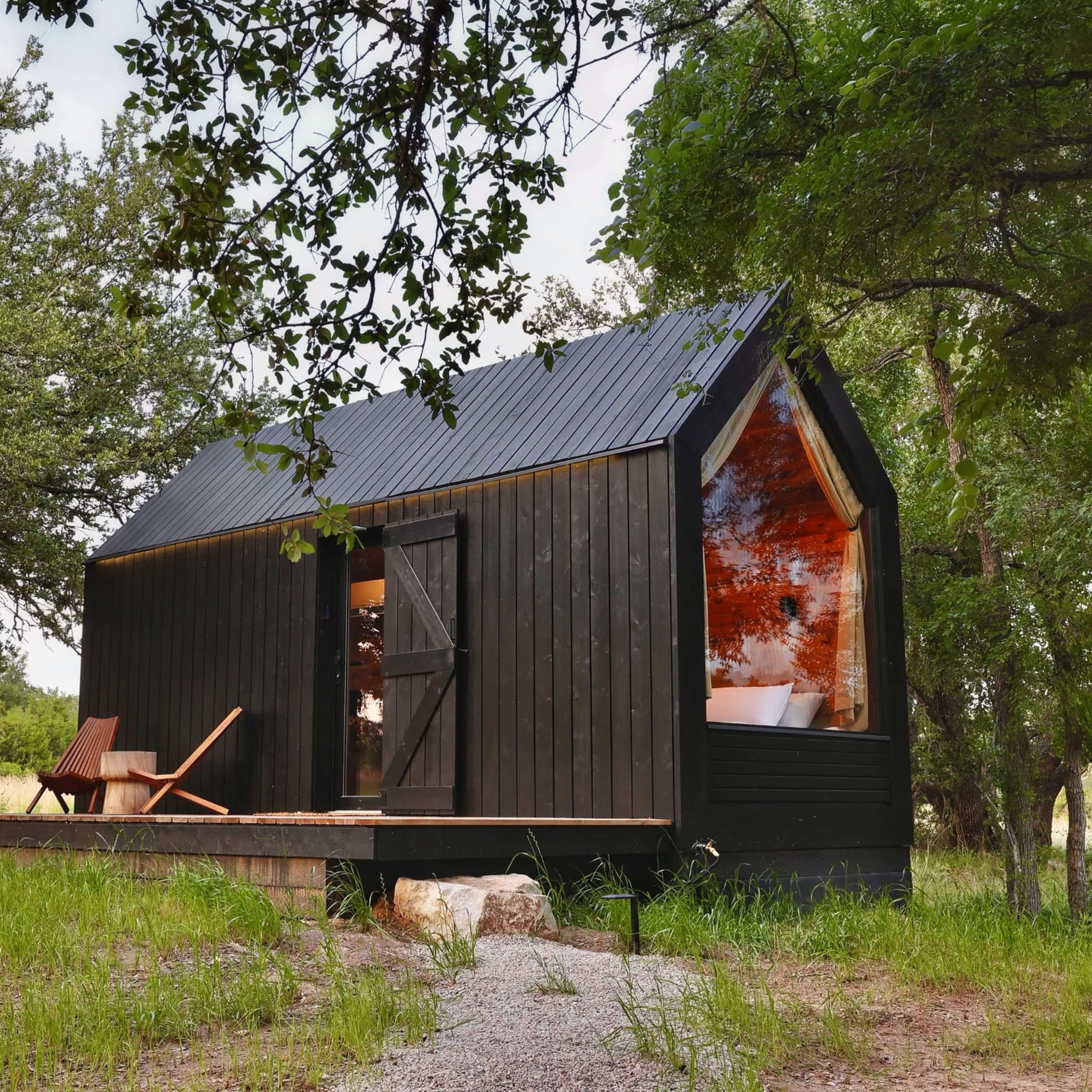 A small black wooden cabin with a metal roof sits surrounded by grass and trees, featuring large windows that reflect the landscape.