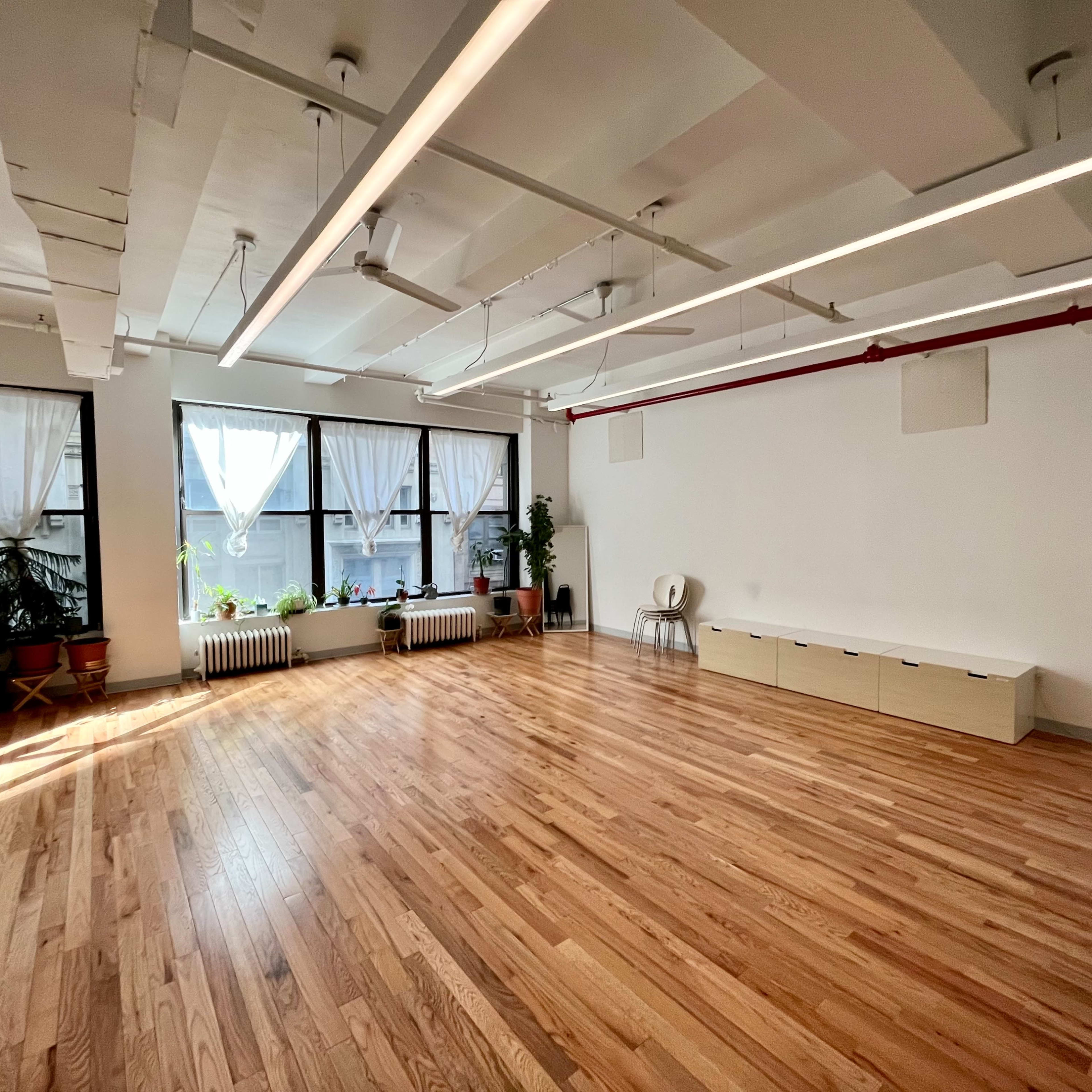 A spacious, well-lit room with hardwood floors, large windows dressed in sheer curtains, and several potted plants.