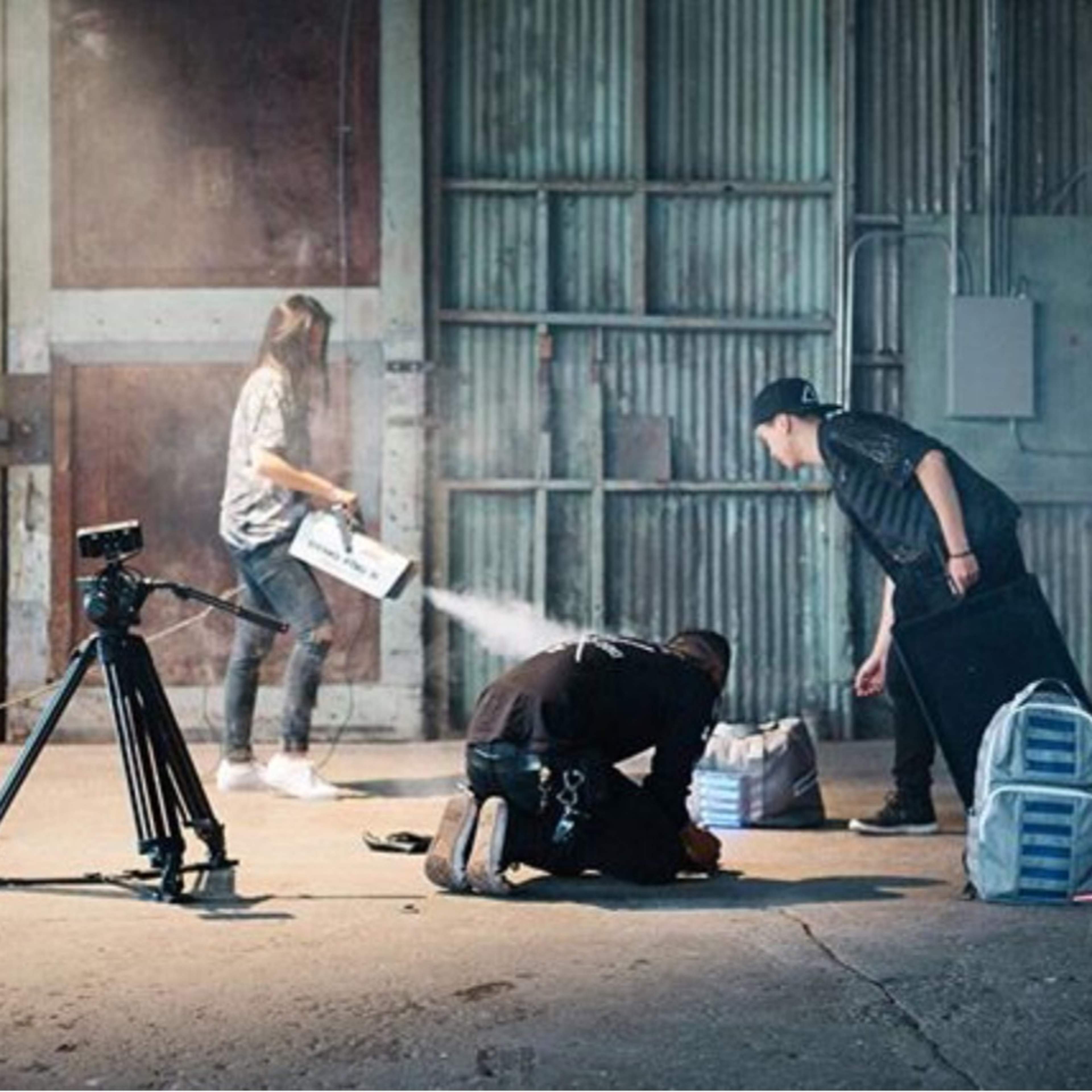 In an industrial setting, two individuals are engaged in tasks near a camera tripod while a third person carries an object in the background.